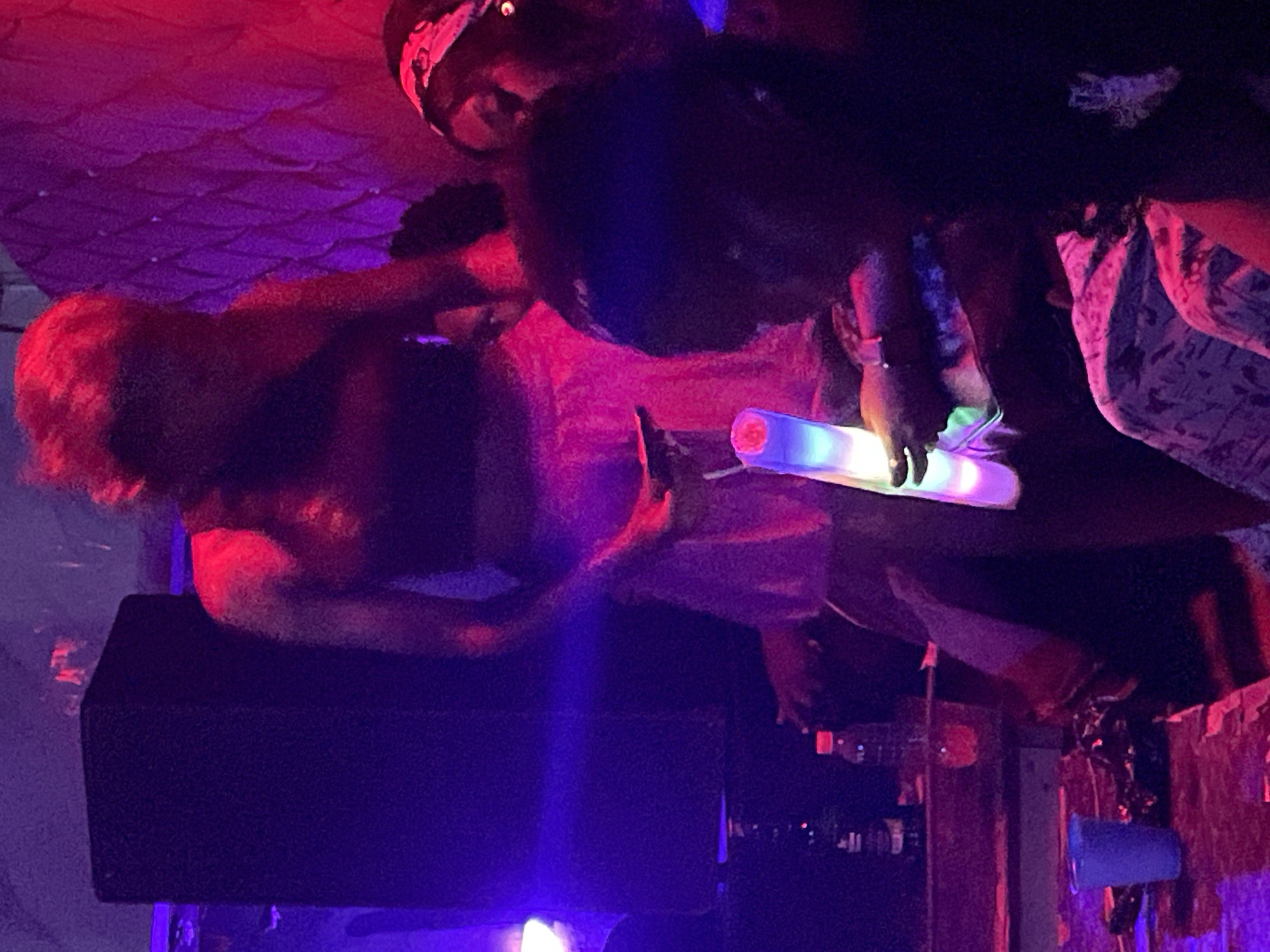 The image depicts a dimly lit indoor scene, likely a nightclub or event space, with vibrant purple and red lighting illuminating the environment. In the foreground, a person's upper body is visible from the chest up, with their head out of frame. They are holding a multi-colored LED foam stick in their right hand, and their left hand is partially visible, appearing to be holding a smartphone. Behind this person, a large black speaker is positioned to the left, bathed in purple light. To the right and slightly behind, another individual's face is visible, looking towards the right. They appear to be wearing a headscarf or bandana. In the middle ground, a person with red hair is visible from the chest up, their face obscured by shadow and the lighting. They are wearing a dark, sleeveless top. On a surface in the lower left of the image, a blue plastic cup and some crumpled materials are present, along with a few bottles. The overall ambiance suggests a lively, social gathering. The image does not contain any specific indicators that would allow for identification of the location within the city of Durimi, Nigeria.