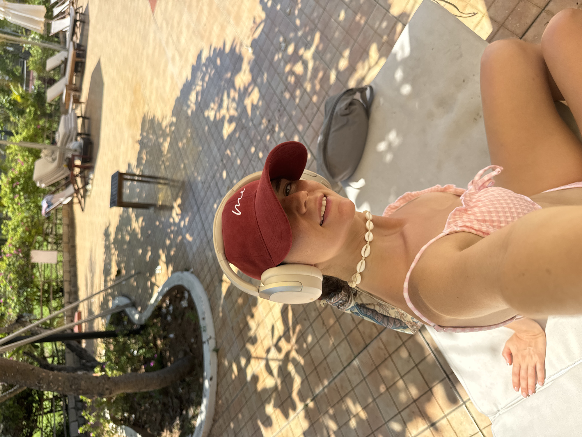 The image shows a woman lying on a lounge chair outdoors, seemingly relaxing. She is wearing a pink gingham bikini top, a maroon baseball cap, and a seashell necklace. She has on beige headphones and is smiling at the camera. The scene is set on a tiled patio with dappled sunlight filtering through trees. In the background, there are more lounge chairs, some tables, and lush green foliage. A grey bag is placed on the patio near her. The time of day appears to be daytime, with bright sunlight. The setting suggests a resort or a place with an outdoor leisure area, possibly a poolside or garden. The woman's expression and attire suggest a relaxed and happy mood. There is no visible text in the image.