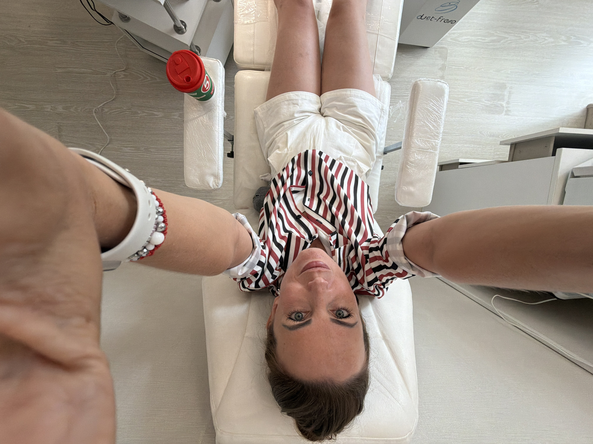 A woman with brown hair pulled up in a bun, wearing a red, white, and black striped shirt and white shorts, lies on a white examination table. Her arms are outstretched as if taking a selfie. She is wearing a white smartwatch with a red and silver beaded bracelet on her left wrist. A red and green cup sits on a white cushion next to the table on the right. The room appears to be a medical or spa setting with light-colored wooden flooring. There is no indication of the city or country.