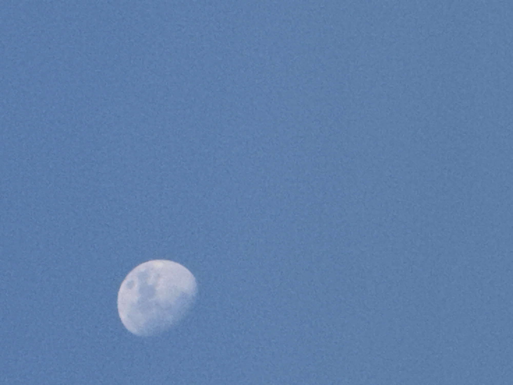 This is a photograph of the moon in a clear blue sky, taken during the daytime. The moon appears as a partially illuminated orb, showing some of its surface features and craters. The sky is a uniform light blue, indicating fair weather and daylight. There is no other discernible activity, objects, or people visible in the frame, nor any text. The setting appears to be an open outdoor space with an unobstructed view of the sky. The image is likely taken from Budva, Montenegro, as per the location context. The time of day is clearly daytime, as indicated by the blue sky and the visible moon. The overall mood is serene and contemplative, focusing on the celestial body against the expansive sky.