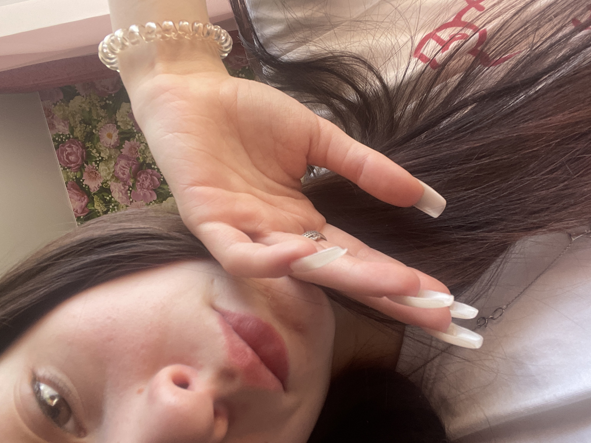 A close-up, tilted shot captures a person partially obscured by their hand, lying down in an indoor setting. The image is taken from an upward angle, showing a portion of the person's face, their hand, and dark hair.
The subject has long, dark brown hair, which is spread out in the frame. Their left hand, featuring long, light-colored (likely white or off-white) acrylic or gel nails, is positioned across their face, covering the forehead and one eye. A silver-toned ring with small stones is visible on one of the fingers. Around the wrist, a clear, spiral-shaped hair tie is worn as a bracelet. The visible part of the person's face includes one eye looking towards the camera, a portion of the nose, and lips painted with a soft pink or muted red lipstick. A silver necklace chain with a clasp is visible near the neck, resting on a white fabric surface.
The background elements include a floral pattern with pink, white, and green flowers, possibly wallpaper or a book cover, seen behind the person's wrist. The hair rests on a white or light-colored fabric, which exhibits a faint, repeating red pattern that resembles stylized letters or symbols. The overall scene suggests a casual pose, possibly a selfie, within a personal space. The lighting is soft. The contextual location provided is Медведево, Russia. No clear, readable text is visible in the image.