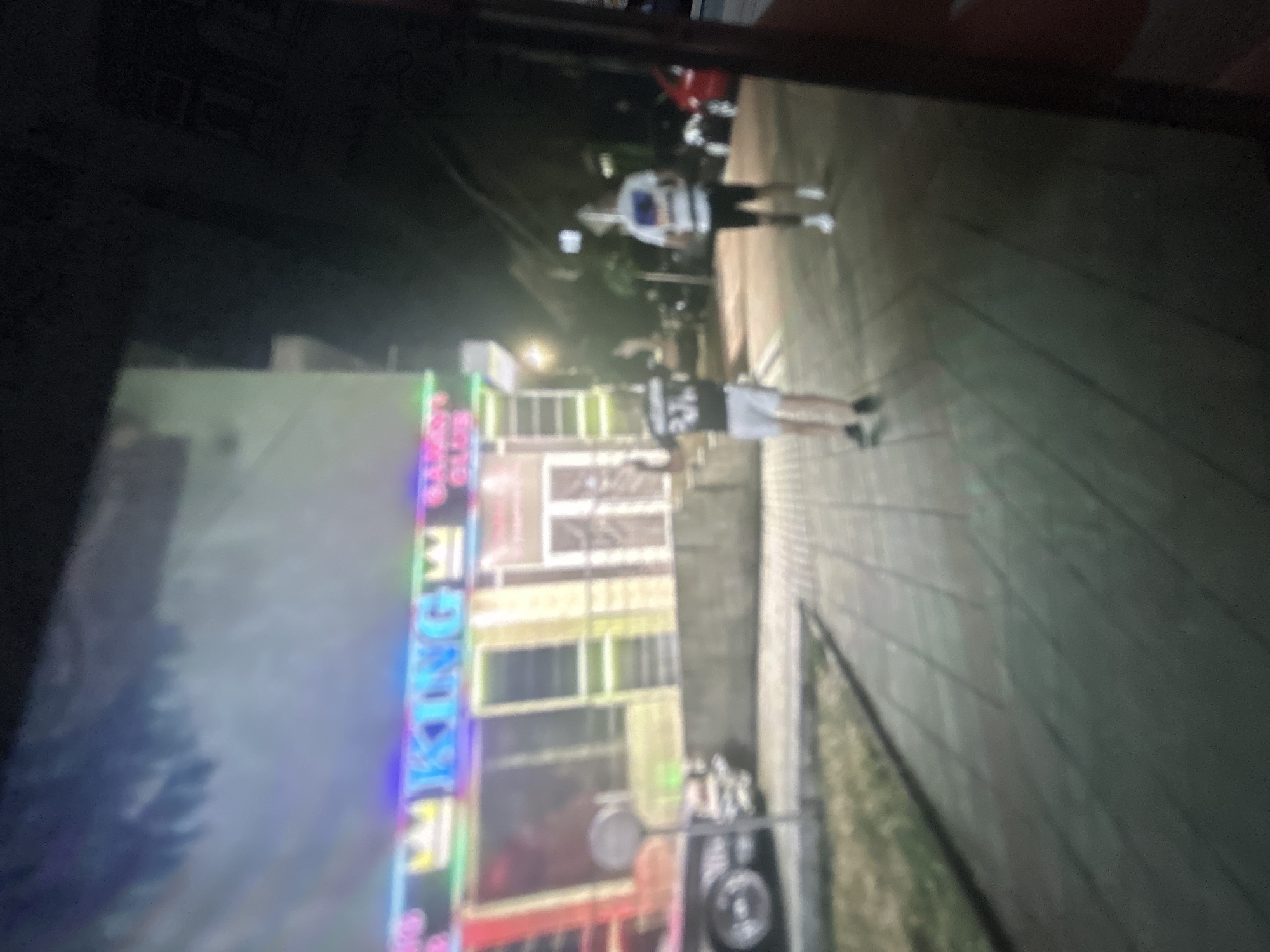 The image shows a nighttime scene on a city street. On the left, there is a building with a brightly lit neon sign that reads "KING CLUB". In the foreground, two individuals are walking along a tiled sidewalk, away from the camera. The first individual is wearing a white jersey and dark shorts, and the second individual is wearing a dark jersey and light shorts. The environment includes trees and streetlights. The location is Unknown, Unknown.