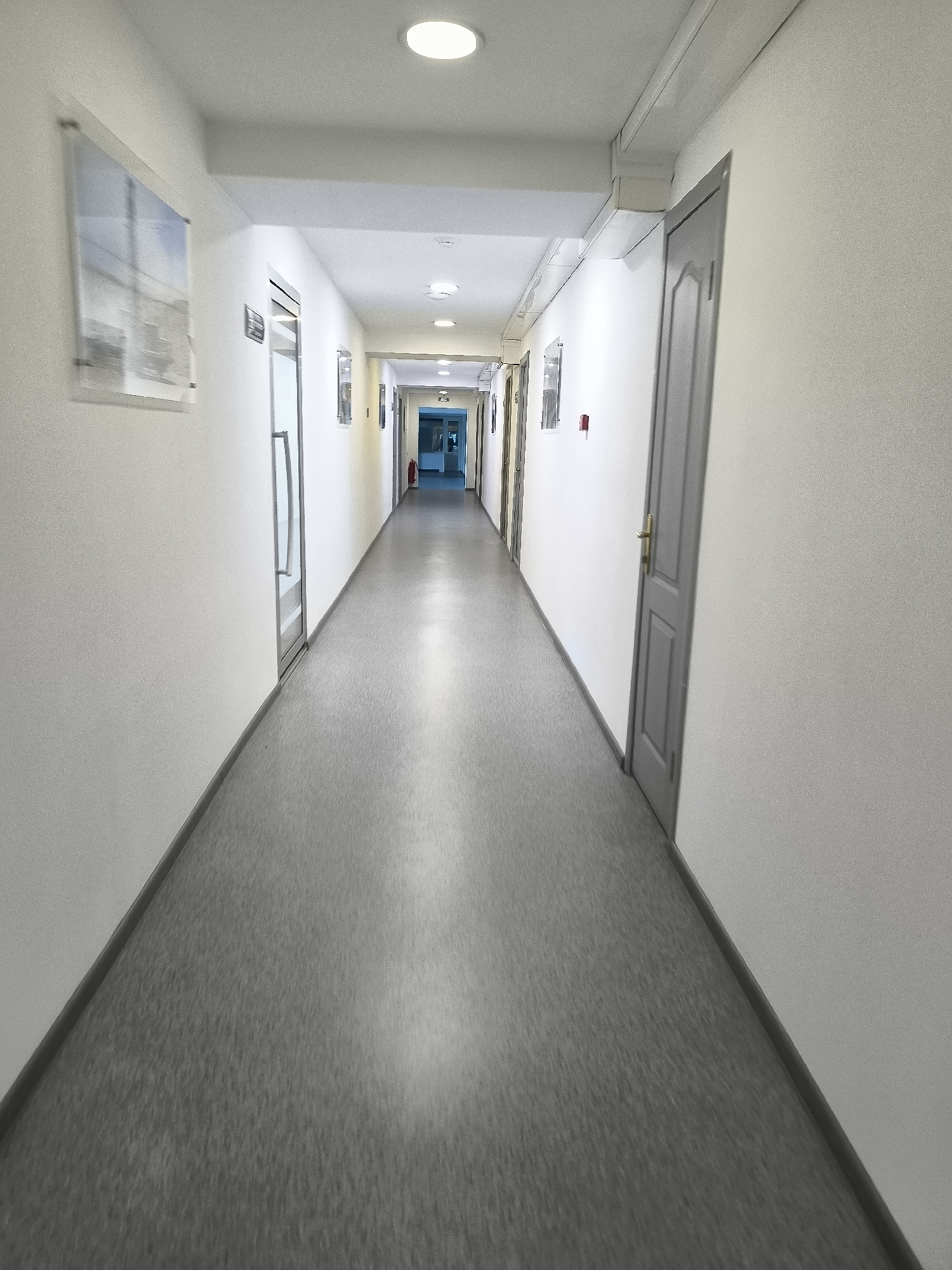 The image shows a long, empty hallway within a building. The hallway has white walls and a grey floor. There are doors on both sides of the hallway, and some are slightly ajar, revealing glimpses of other rooms or spaces beyond. Overhead, there are circular fluorescent lights illuminating the corridor. On the left wall, there is a framed piece of artwork or a poster. Towards the end of the hallway, there are signs on the doors and what appears to be a fire extinguisher mounted on the wall. The hallway extends into the distance and appears to be well-maintained. The scene is static and devoid of any human presence. This location is within the Qarağandı city administration building in Qarağandı, Kazakhstan.