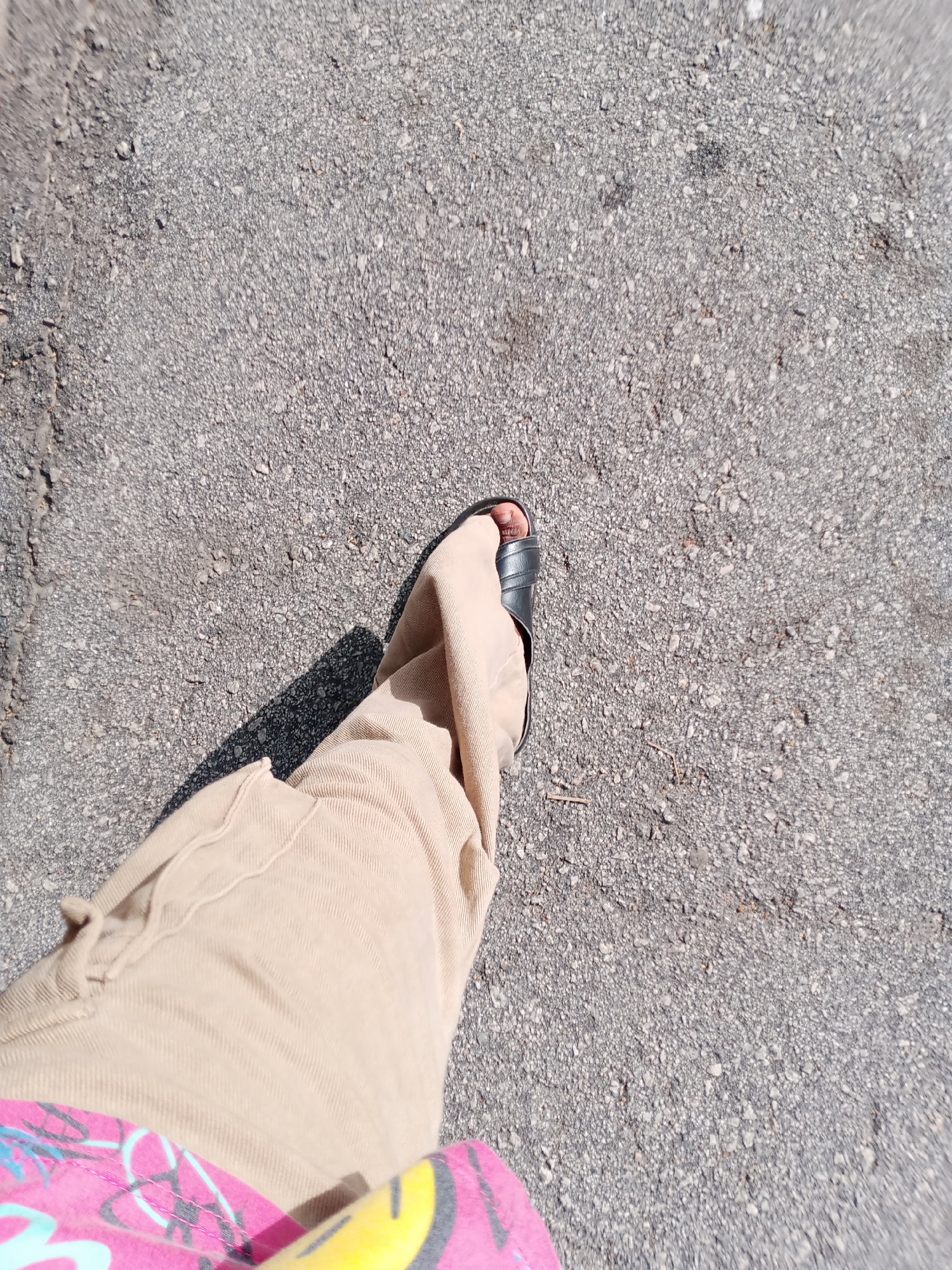 The image shows a close-up, high-angle view of a person's legs and feet walking on an asphalt surface. The person is wearing tan-colored pants and black sandals. A colorful pink, blue, and yellow patterned fabric is visible at the bottom of the frame, partially obscured by the pants. The asphalt is gray and textured, with small stones and cracks visible. A shadow is cast to the left of the person's right leg. The image was taken outdoors, likely on a sunny day, given the shadow. There is no information in the image to suggest the location is Jalingo, Nigeria.