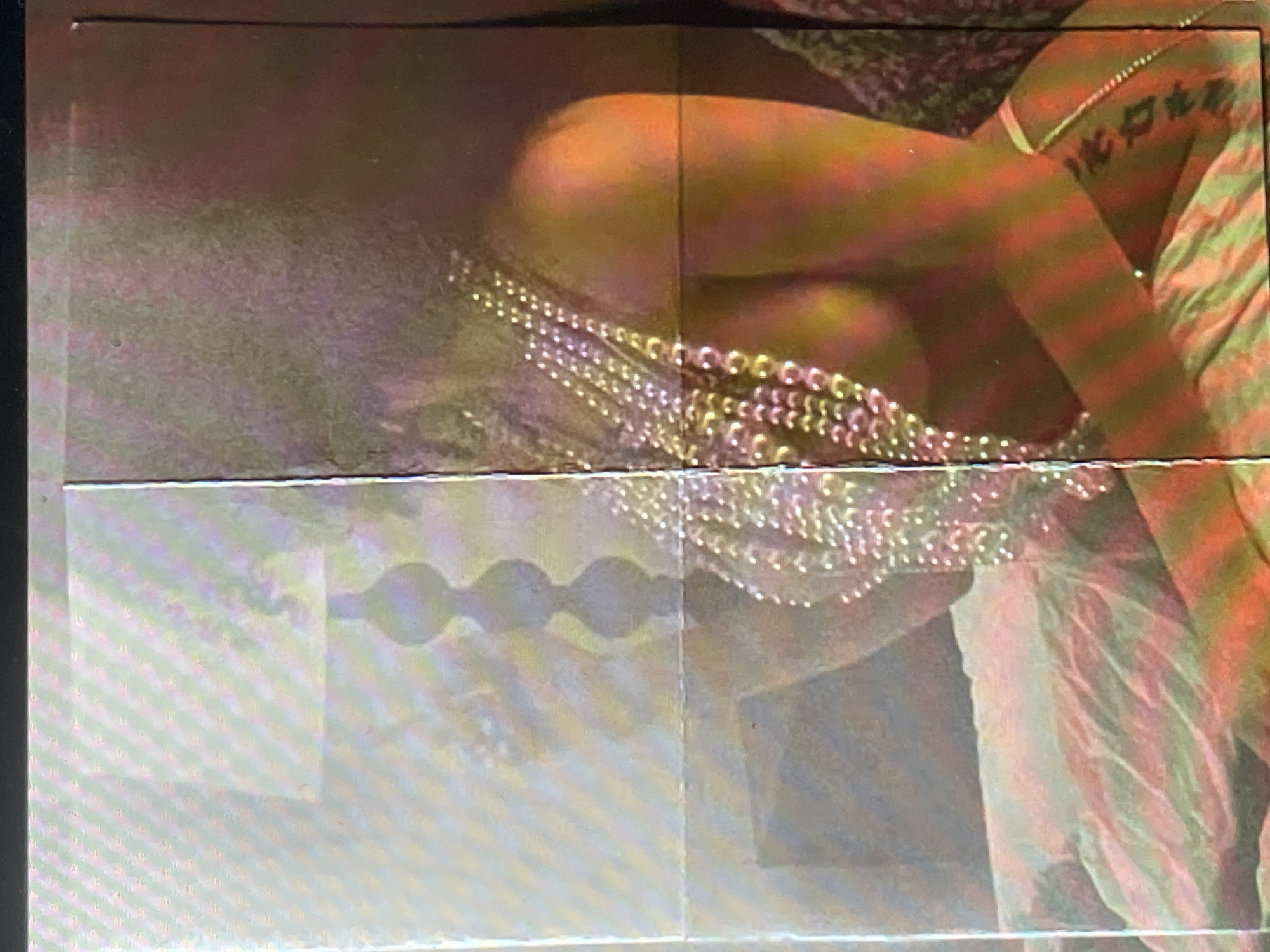 This media file presents a close-up, artificially lit view of a person, likely performing or posing, within an indoor setting. The image focuses on the upper body, encompassing the torso, arm, and hip area. The individual is adorned with numerous strands of glistening, light-colored beads or chains, draped across their midsection, creating a shimmering effect under the dramatic lighting.
The scene is characterized by vibrant, warm, artificial lighting, primarily in shades of orange, red, and yellow, which strongly illuminate the person's skin and the beads. On the right side, a flowing fabric, possibly part of a costume or stage drapery, is visible with distinct vertical stripes in hues of red, orange, and muted green. A clear tattoo, featuring two Chinese characters, "龍" (lóng - dragon) and "虎" (hǔ - tiger), is visible on the person's upper arm.
In the lower section of the image, particularly towards the left, there is a visible pattern of connected, wavy, or circular shapes against a lighter, somewhat grainy background. This might be a reflection, a projected image on a screen, or part of the stage design. The overall image displays signs of digital artifacts, including prominent horizontal lines or bands, general graininess, and a slight lack of sharp focus, suggesting it may be a photograph of a screen or a frame from a video. The context implies an event such as a performance, concert, or photoshoot, possibly in a venue located in Sofia, Bulgaria, although the image itself contains no specific geographical identifiers beyond the general setting.