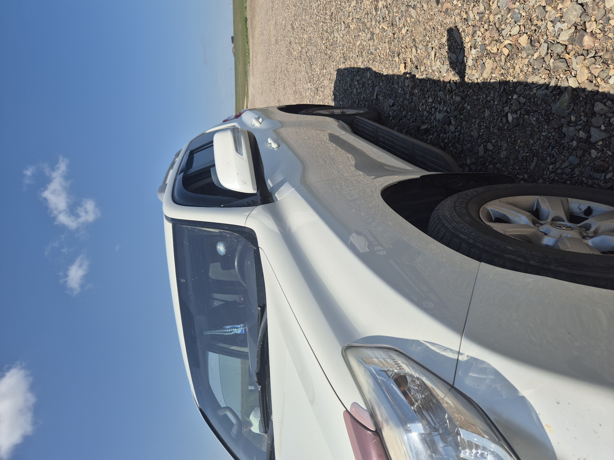 A white SUV is parked on a gravel road with a wide, open grassy plain in the background. The sky is bright blue with a few scattered white clouds. The car is angled towards the viewer from the front left, showing its headlight, hood, windshield, side mirror, and part of the passenger door and running board. The terrain suggests a rural or undeveloped area. There is a person visible inside the car through the windshield. In the distance, on the horizon, there are some structures that appear to be buildings or other man-made objects, possibly associated with agriculture or rural living, in the vicinity of Novo-Ishim Rural District, Kazakhstan.