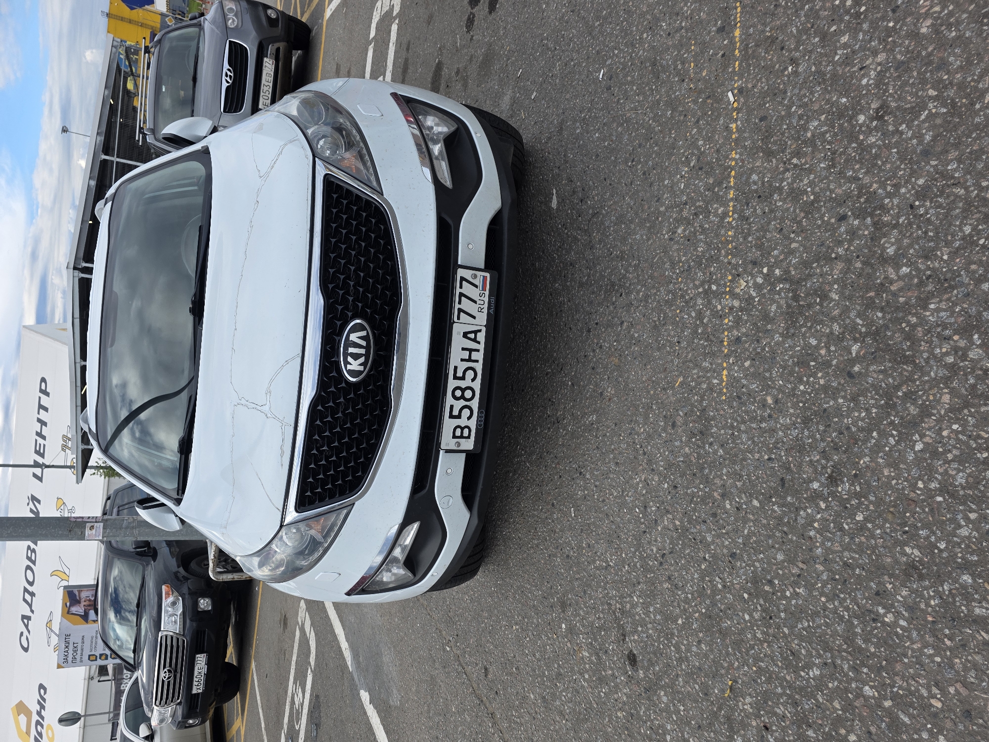 This media file shows a front-end close-up view of a white Kia Sportage parked on asphalt. The car is the primary subject, with its grille, headlights, and license plate prominently displayed. The license plate reads "B585HA 777 RUS". To the left of the Kia, a dark-colored SUV, possibly a Toyota Land Cruiser, is partially visible, also parked. Its license plate reads "X660KE 777". A grey Hyundai Santa Fe is visible behind the Kia, with its license plate reading "E053 EB 27".
In the background, there are signs with text in Russian. One sign on the left reads "САДОВЫЙ ЦЕНТР" (Gardening Center). Another sign below it has text including "ЗАКАЖИТЕ ПРОЕКТ ДЛЯ ВАШЕГО ДОМА" (Order a project for your home) and "БЕСПЛАТНО СПРОЕКТИРУЕМ" (We will design for free). The sky is visible in the upper left corner, with clouds, suggesting it is daytime. The setting appears to be a parking lot, likely at a commercial establishment related to home and garden, given the signage. There are no people visible in the image. The overall impression is a snapshot of cars parked in an outdoor commercial area.