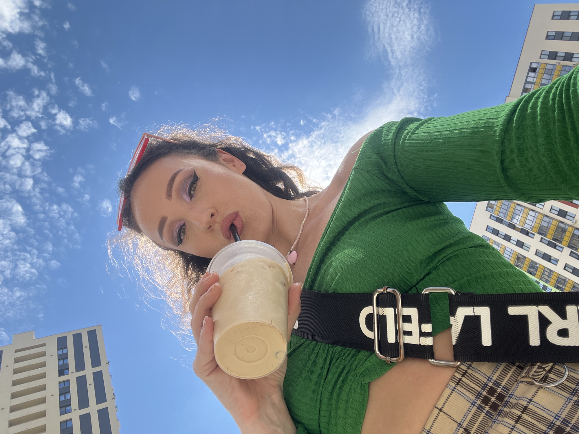 A low-angle, wide shot captures a woman drinking from a disposable cup while outdoors. She is wearing a green off-the-shoulder top and a plaid skirt. A black strap with white lettering, reading "KARL LAGERFELD", is visible across her chest. Red sunglasses are perched on her head. In the background, two modern apartment buildings are visible against a bright blue sky with wispy clouds. The scene appears to be set in a city, and considering the specific architectural style of the buildings, the location is consistent with the city of Kazan, Russia.