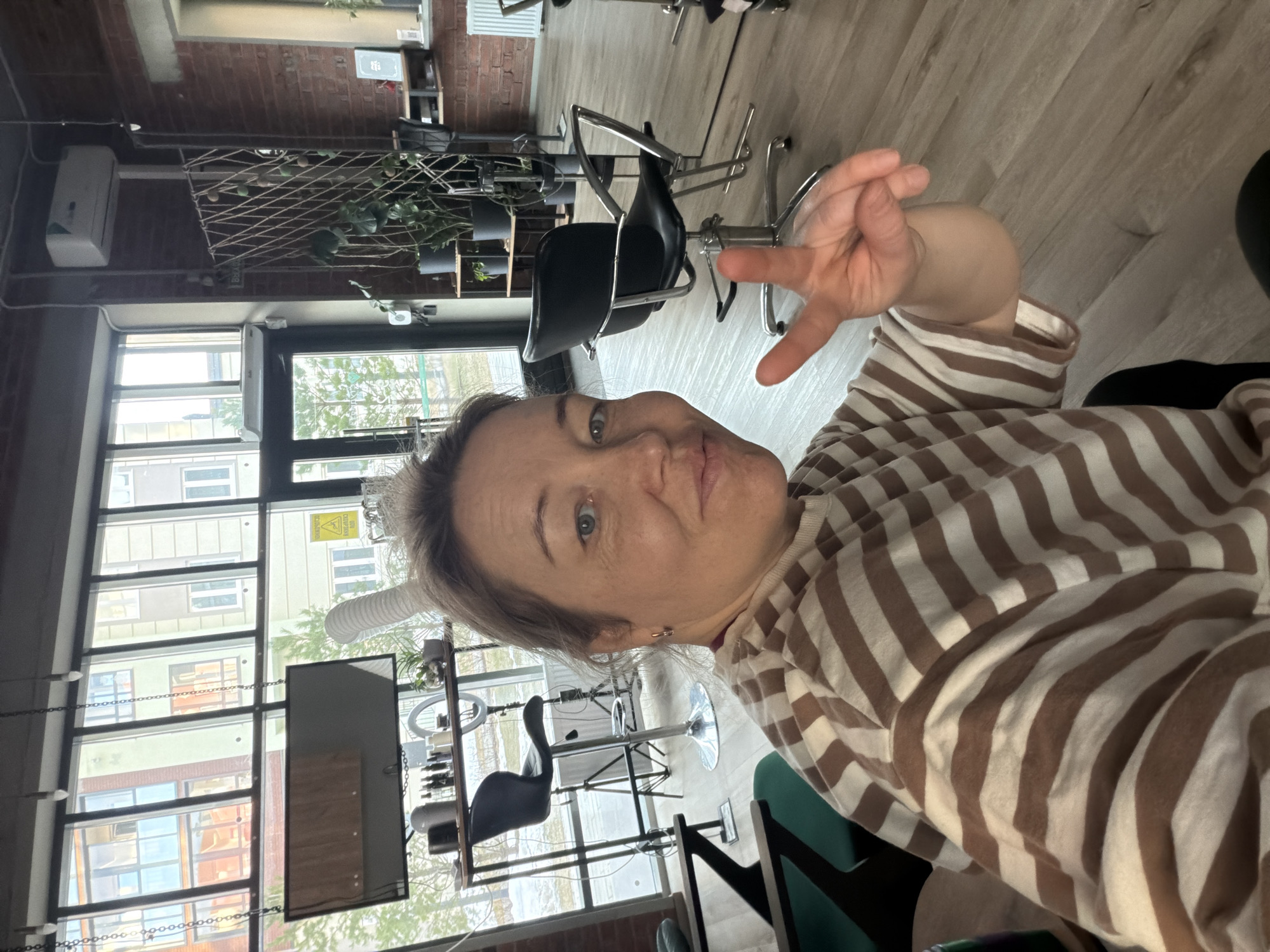 A woman is taking a selfie indoors in Pervomaysky, Russia, likely at a hair salon. She is wearing a brown and white striped shirt and making a hand gesture. Behind her are hair salon chairs and windows showing buildings across the street. There is salon equipment visible, including a ring light and a mirror. The interior has a modern industrial style with exposed brick.
