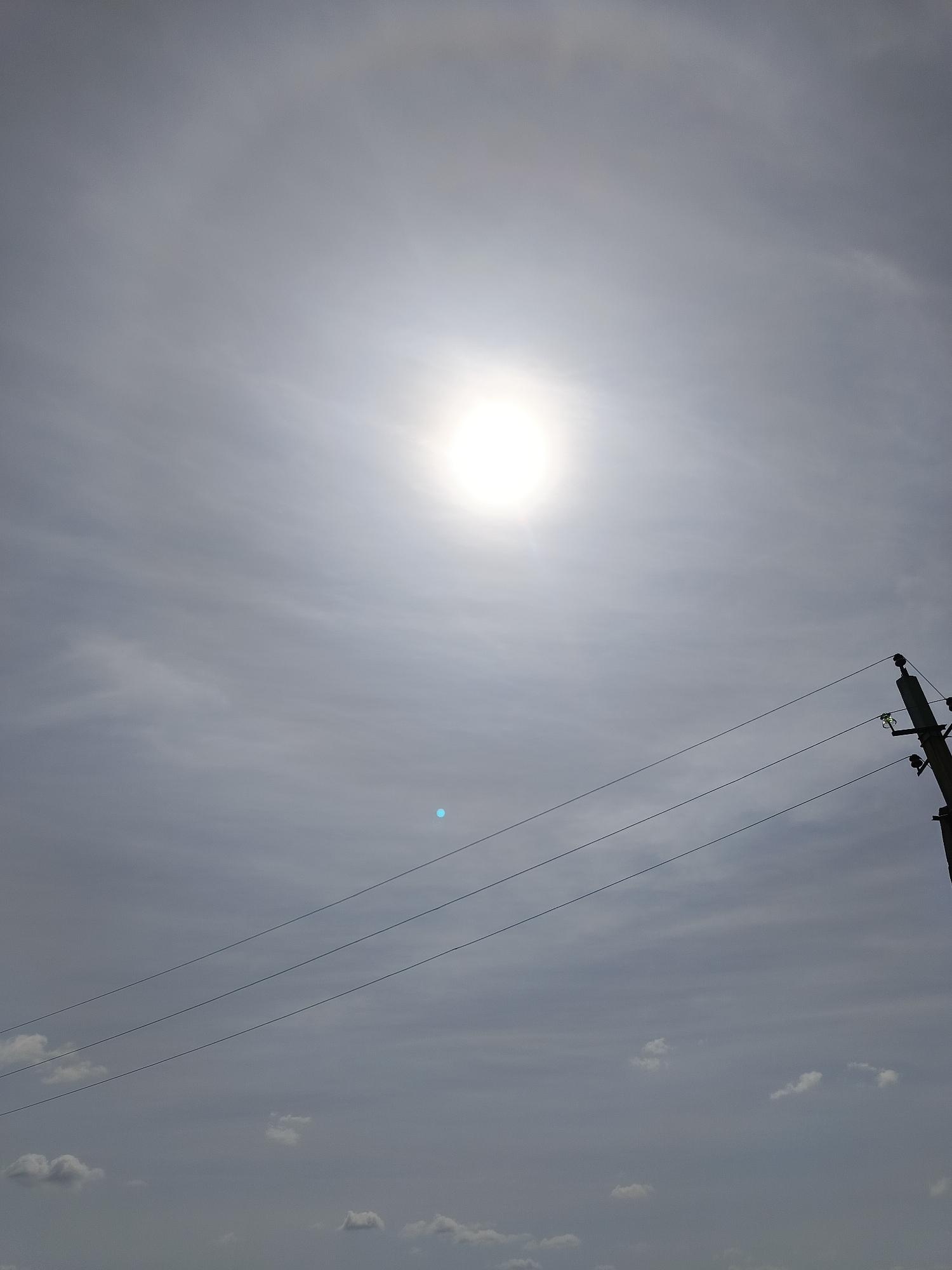 The image shows a daytime sky with a bright sun encircled by a halo, likely a 22° halo. The sun is high in the sky, slightly off-center. Power lines run diagonally across the lower right portion of the image, attached to a utility pole on the right edge. A few scattered clouds are visible in the lower part of the image. Based on the power lines and cloud formations, and the presence of the halo, the location is identified as Chaikovsky, Russia. No people or animals are visible.