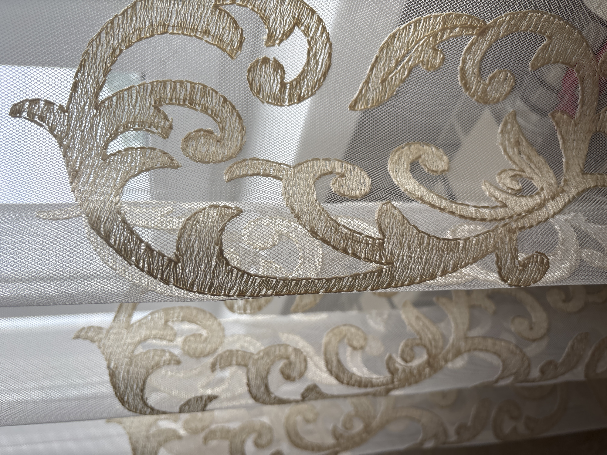 This is a close-up, high-angle, full-shot photograph of sheer, beige, embroidered curtains. The curtains are made of a fine mesh fabric with a repeating pattern of gold thread embroidery in a scrolling, floral, Baroque style. The embroidery is denser and more prominent on the right side of the image, creating a rich texture against the sheer fabric. Light filters through the curtains from the right, casting shadows and highlighting the intricate details of the embroidery. The background is out of focus, showing a blurred suggestion of a window with light beyond. The scene is indoors, likely a domestic setting. There is no indication of the city of Bila Tserkva, Ukraine.
