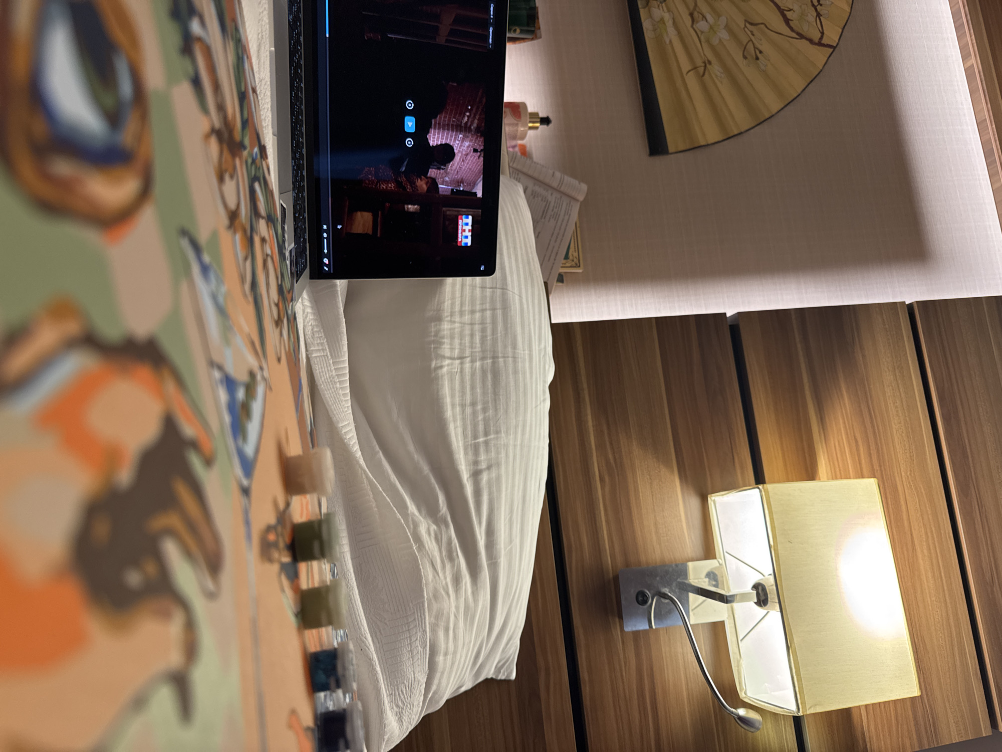 The image depicts a bedside scene with a laptop displaying a video, a pillow, and a bedside lamp. A decorative fan is visible on the left, and a close-up of what appears to be a paint-by-numbers kit with small paint pots is in the foreground. The wall behind the bed has horizontal wooden paneling, and a wall lamp with a light-yellow shade is illuminated. The scene is set in a bedroom. There is no visual information within the image to suggest a location in Aksu, Turkey.