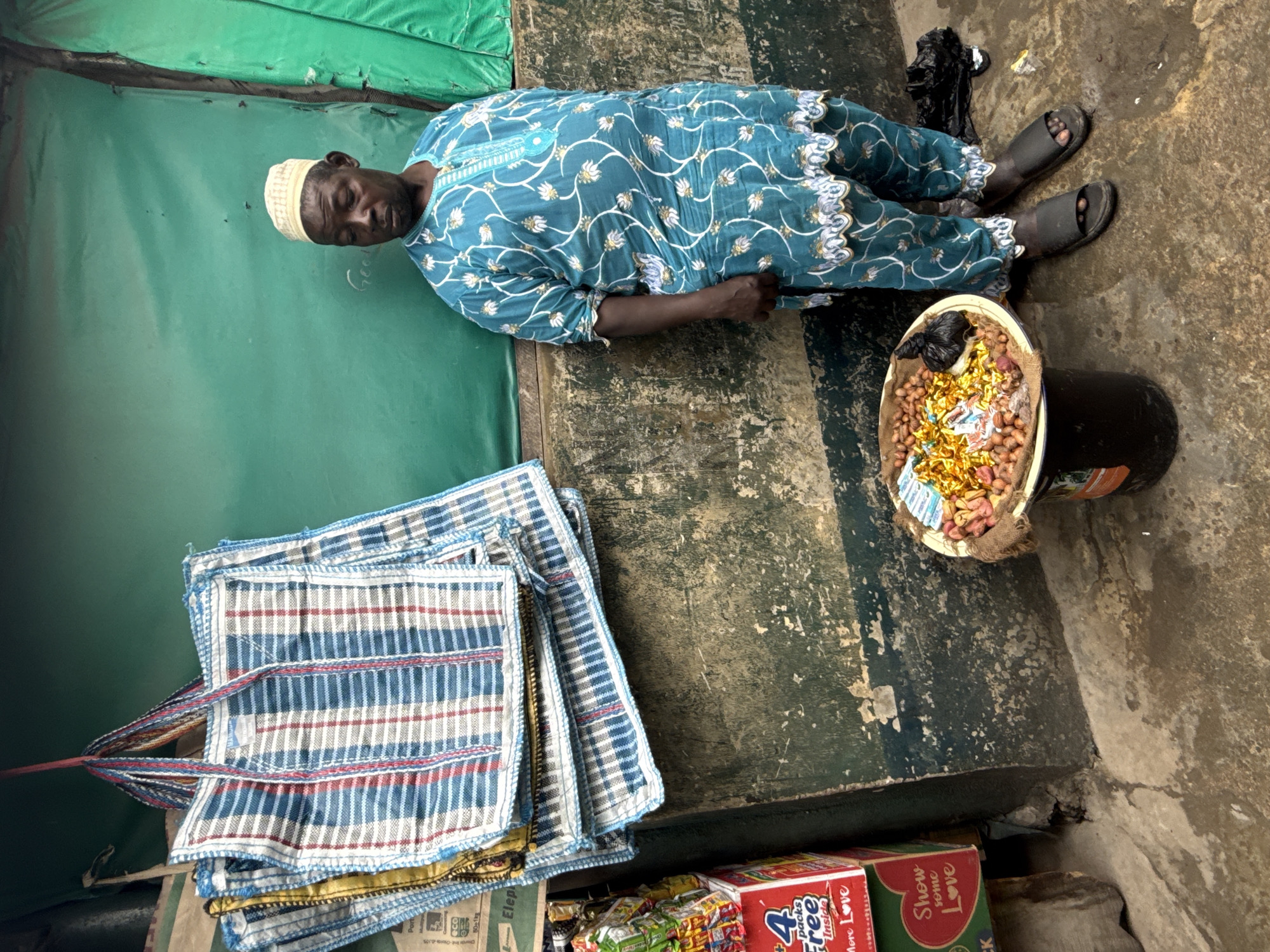 An adult male, wearing a teal-colored kaftan with a white floral pattern and a white cap, stands in front of a green tarp backdrop in Lagos, Nigeria. He is positioned on the right side of the image, looking to his left. In front of him, a black bucket holds a round, shallow bowl overflowing with small, brown nuts, some wrapped in gold foil. Beside the bucket, a black plastic bag lies on the wet ground. To the left of the man, several large, plaid tote bags, predominantly blue and white with red stripes, are stacked and hanging from a red string. In the lower left corner of the frame, a red cardboard box with a white and green design is visible, displaying the text "4 packs Free Inside" and "More Love". The ground is wet and cracked, suggesting it may have recently rained. The wall behind the man is textured and appears aged, with patches of green and dark paint.