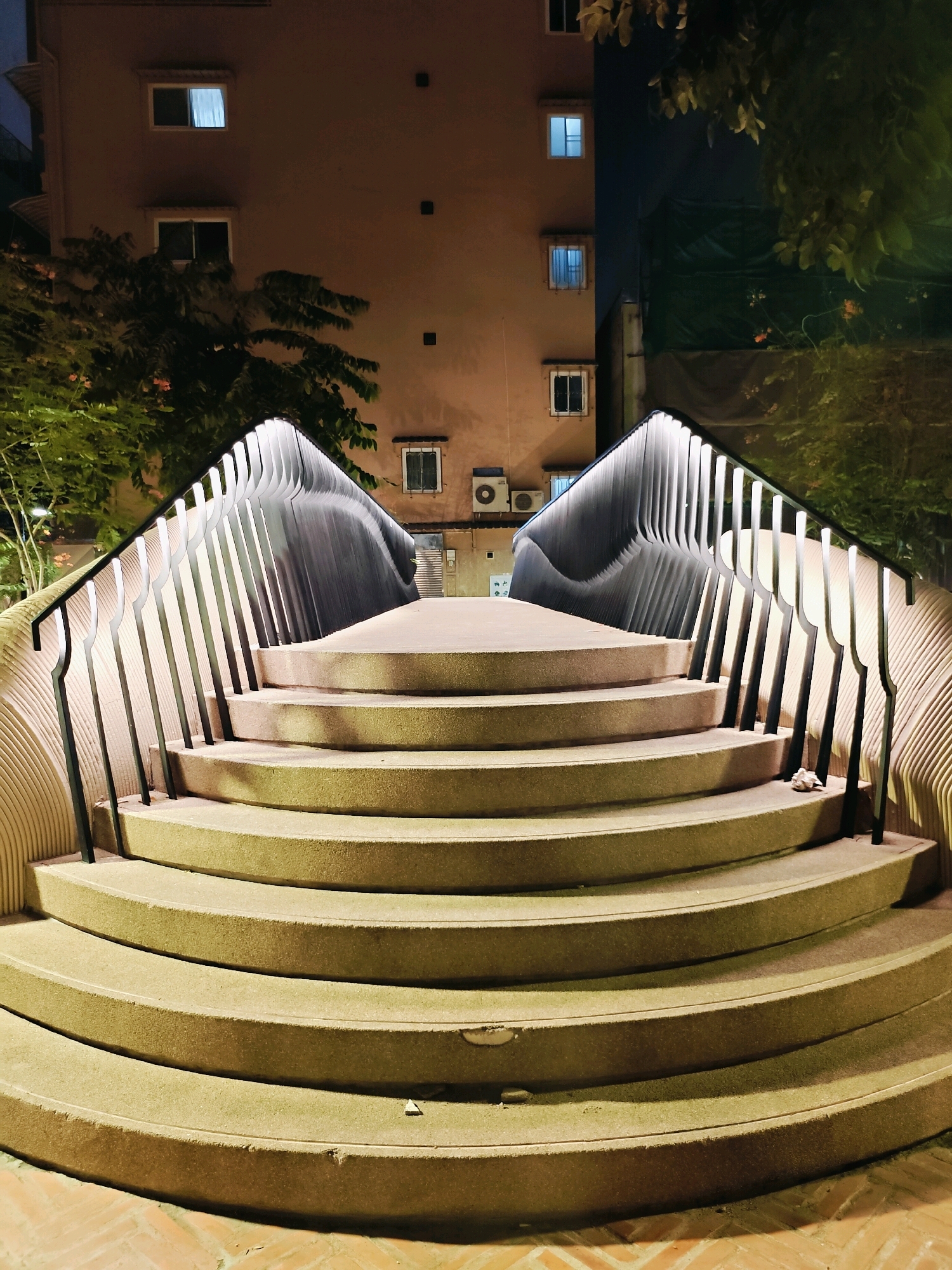 This photograph captures a perspective of a curved, multi-tiered concrete staircase with a modern, illuminated railing, likely taken at night. The staircase is the central focus, with its textured, light-colored steps forming a wide arc. The railing is composed of dark, vertical metal bars interspersed with glowing lights that create a dramatic effect.
Behind the staircase, a building with multiple windows is visible. Some windows are lit from within, casting a faint glow, while others appear dark. The building's facade is a muted, light brown color. Trees with dark green foliage are present on either side of the staircase and the building, with some branches reaching towards the foreground. One tree on the left side appears to have small pink flowers.
To the right of the building, a dark green tarp or curtain is visible, suggesting some form of construction or temporary covering. The ground in the foreground is paved with reddish-brown rectangular tiles arranged in a herringbone pattern. The overall scene is illuminated by artificial light sources, creating contrasting shadows and highlights, and it appears to be an outdoor public space in Bangkok, Thailand. The image has a slightly fish-eye distortion, making the edges appear curved.