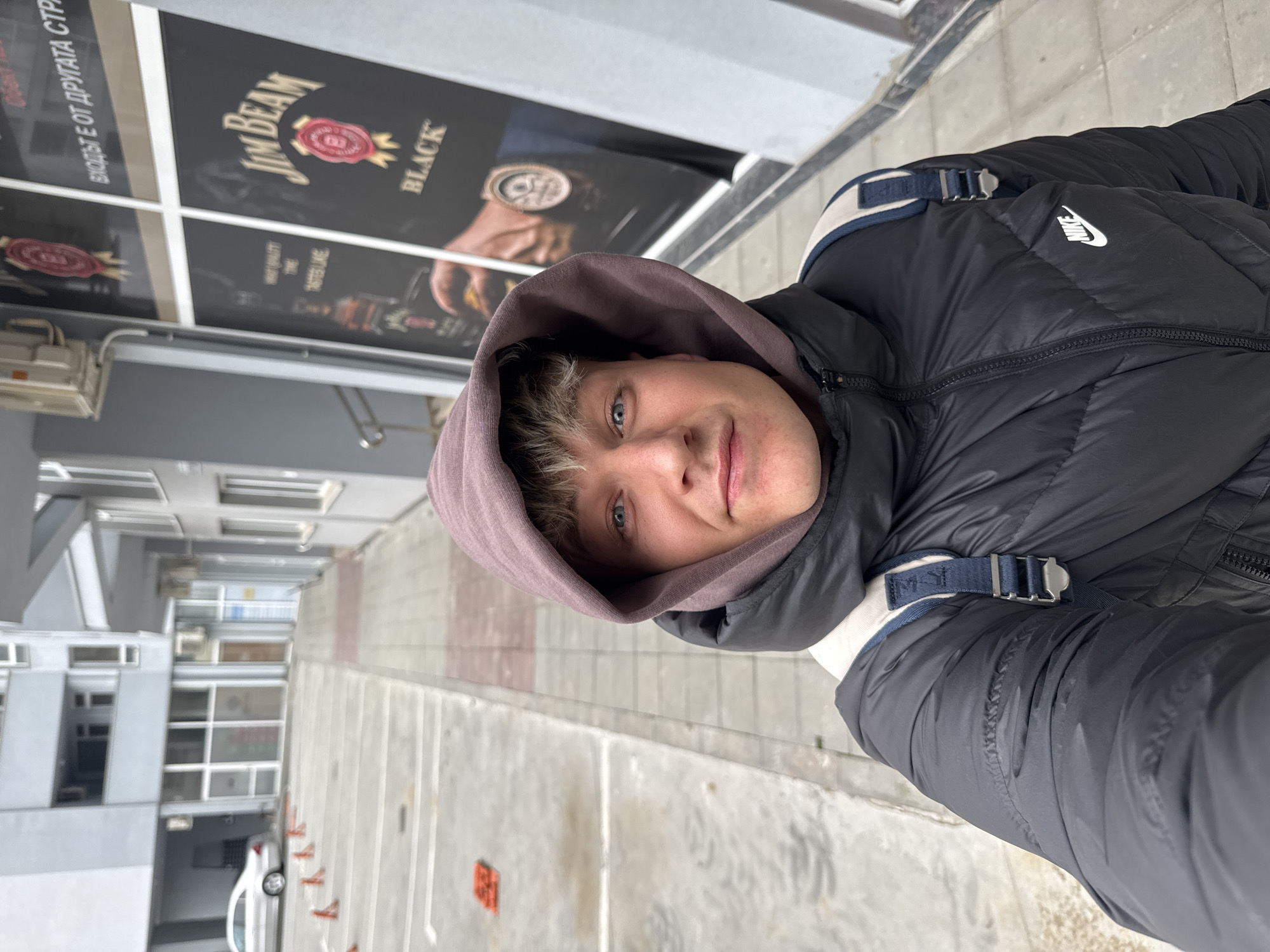 A selfie captures a fair-skinned young man wearing a black Nike puffer jacket with a brown hooded sweatshirt underneath. He gazes directly into the camera, his face partially obscured by the hood. Behind him, a Jim Beam Black advertisement is visible on a building facade. The background features a typical urban street scene with a light-colored building and a narrow roadway marked with orange and white lines. A white car is parked further down the street. The location is Plovdiv, Bulgaria.