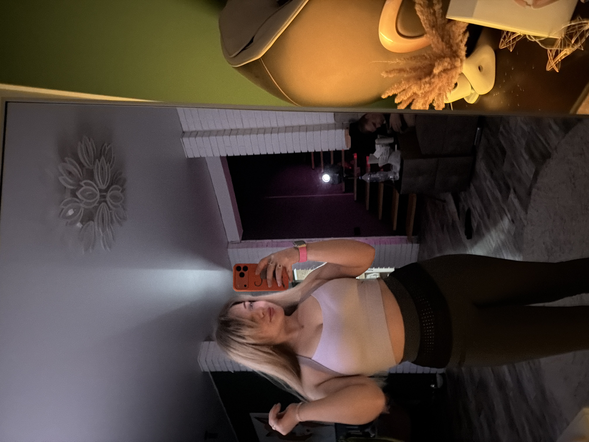 The image shows a reflection in a mirror. A woman with blonde hair, wearing a light-colored sports bra and dark leggings, is taking a selfie with an orange phone. The background visible in the mirror includes a white brick wall, a dark hallway, and a floral wall decoration. Above the mirror and the woman's reflection, there are decorative objects on a surface including a beige hat, a vase with dried flowers, and decorative stars. The location of the image is in Большая Шильна, Russia. The specific details within the hallway scene such as the flooring, furniture, and lighting conditions are not clear enough to provide additional forensic detail.