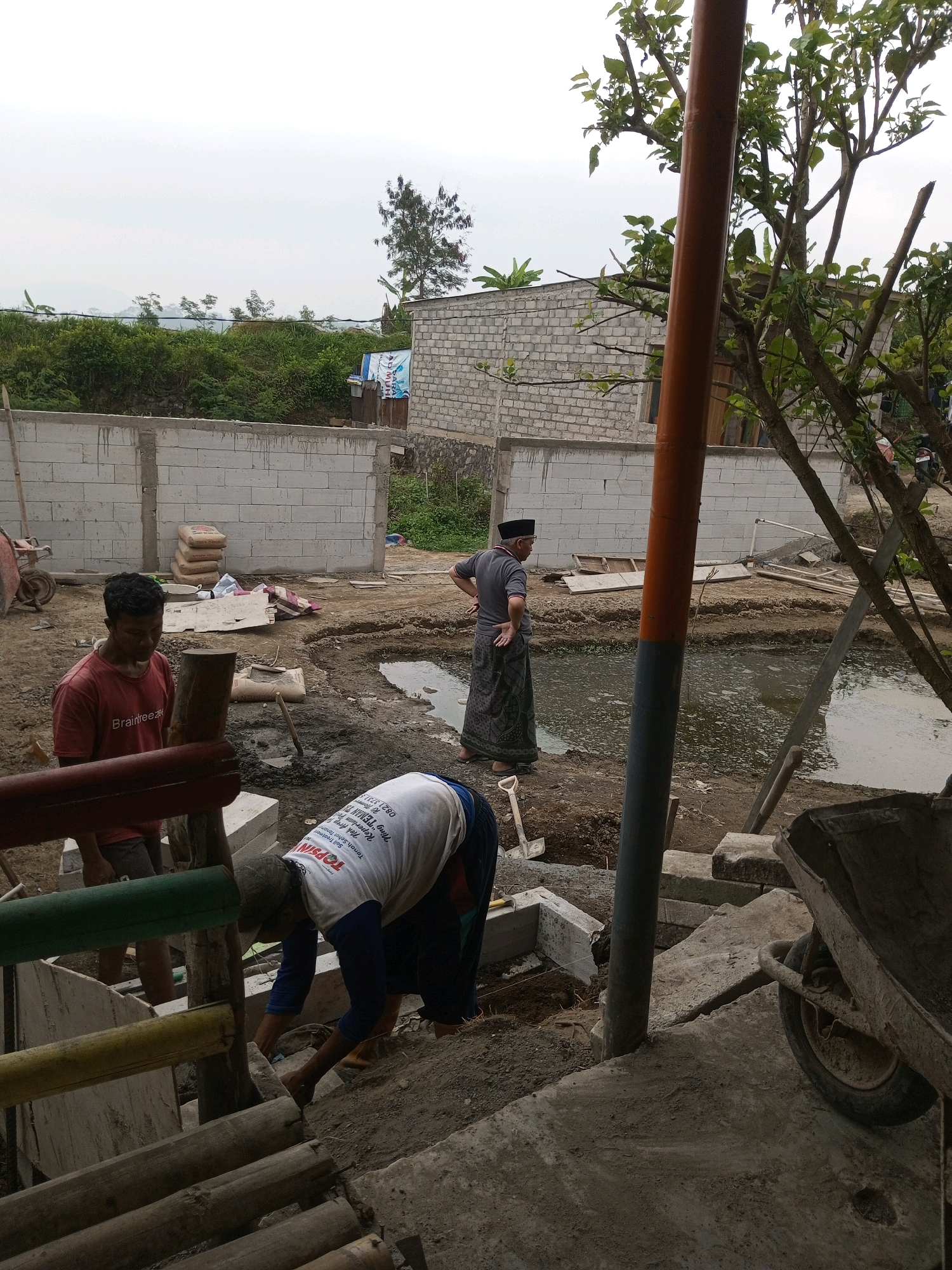 The image depicts an outdoor construction site in the village of Kalibeber, Indonesia. In the foreground, a man in a red t-shirt with "Braintree" printed on it is standing and looking down. To his right, another man wearing a white t-shirt with text that includes "TOPSIN" and a dark blue long-sleeved shirt underneath is bent over, working on the ground. He is wearing dark pants. Further back, a third man in a grey shirt and a black peci cap is standing with his hands on his hips, observing the work. He is wearing a patterned sarong.
The environment is dusty and filled with construction materials. A large pool of murky water is visible in the middle ground. There are stacks of cement bags leaning against a white brick wall in the background. Several lengths of bamboo are visible in the lower foreground, suggesting ongoing construction. A wheelbarrow is positioned to the right of the frame, also covered in dirt. The sky is overcast. The scene appears to be a residential construction project in a rural setting.
