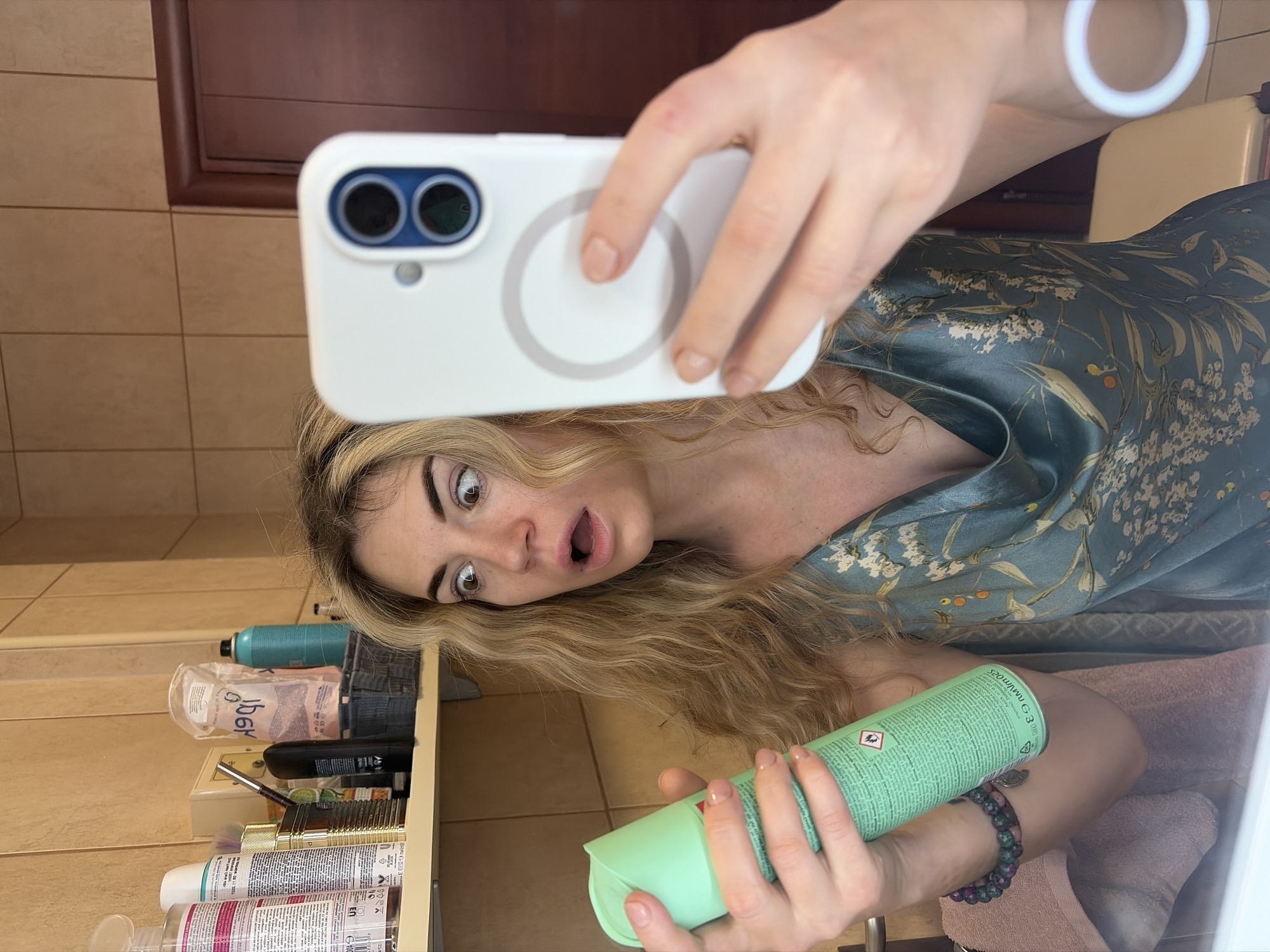 The scene depicts a woman taking a mirror selfie in a bathroom, reportedly located in Budva, Montenegro. The woman, with blonde, curly hair, is positioned centrally in the frame. Her eyes are wide, and her mouth is open, conveying an expression of surprise or shock. She is wearing a blue-green satin top with a light floral pattern.
In her left hand, she holds a light-green aerosol can, partially obscuring text and displaying a red flame hazard symbol. Her left wrist wears a multi-colored beaded bracelet with a metallic charm. In her right hand, she holds a white smartphone with a visible circular MagSafe ring on the back and a dual-camera module featuring blue rings around the lenses. Her fingernails are painted with a light pink polish.
The background consists of light-brown square wall tiles. A white shelf is visible behind the woman, holding various toiletries. These include a white spray bottle with a pink label, a white bottle, a gold-colored container, a black eyeliner pencil, a black deodorant bottle, a clear plastic bag containing white items (possibly cotton pads) with reversed text, a dark grey woven basket, and a teal-colored aerosol can. To the far right, a dark brown wooden door frame is partially visible. The bottom left foreground shows a white surface and a light pink towel, suggesting a sink or counter area.