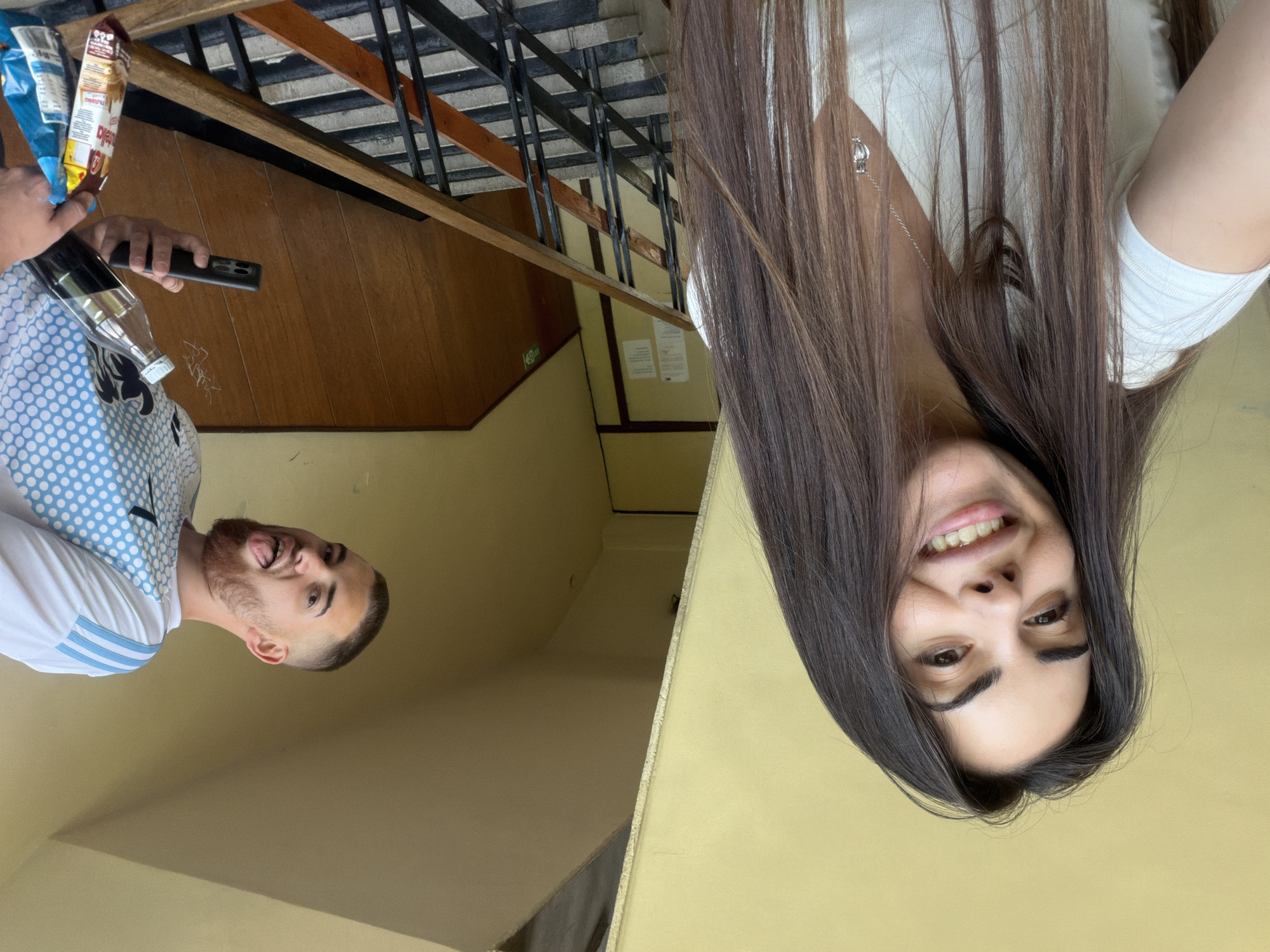 The image captures two individuals in an indoor setting, possibly an apartment building in Sofia, Bulgaria. The foreground features a woman with long, dark brown hair, wearing a white shirt and a silver necklace. She is smiling and appears to be looking at the camera. In the background, a man is holding a smartphone and some snacks (chips) and a glass bottle, possibly taking the picture. He is wearing a white and blue dotted shirt. The background also shows a wooden staircase with black iron railings. The walls are a pale yellow color. The photograph seems to have been taken from a slightly tilted angle. There are some posters or notices hanging on the walls.