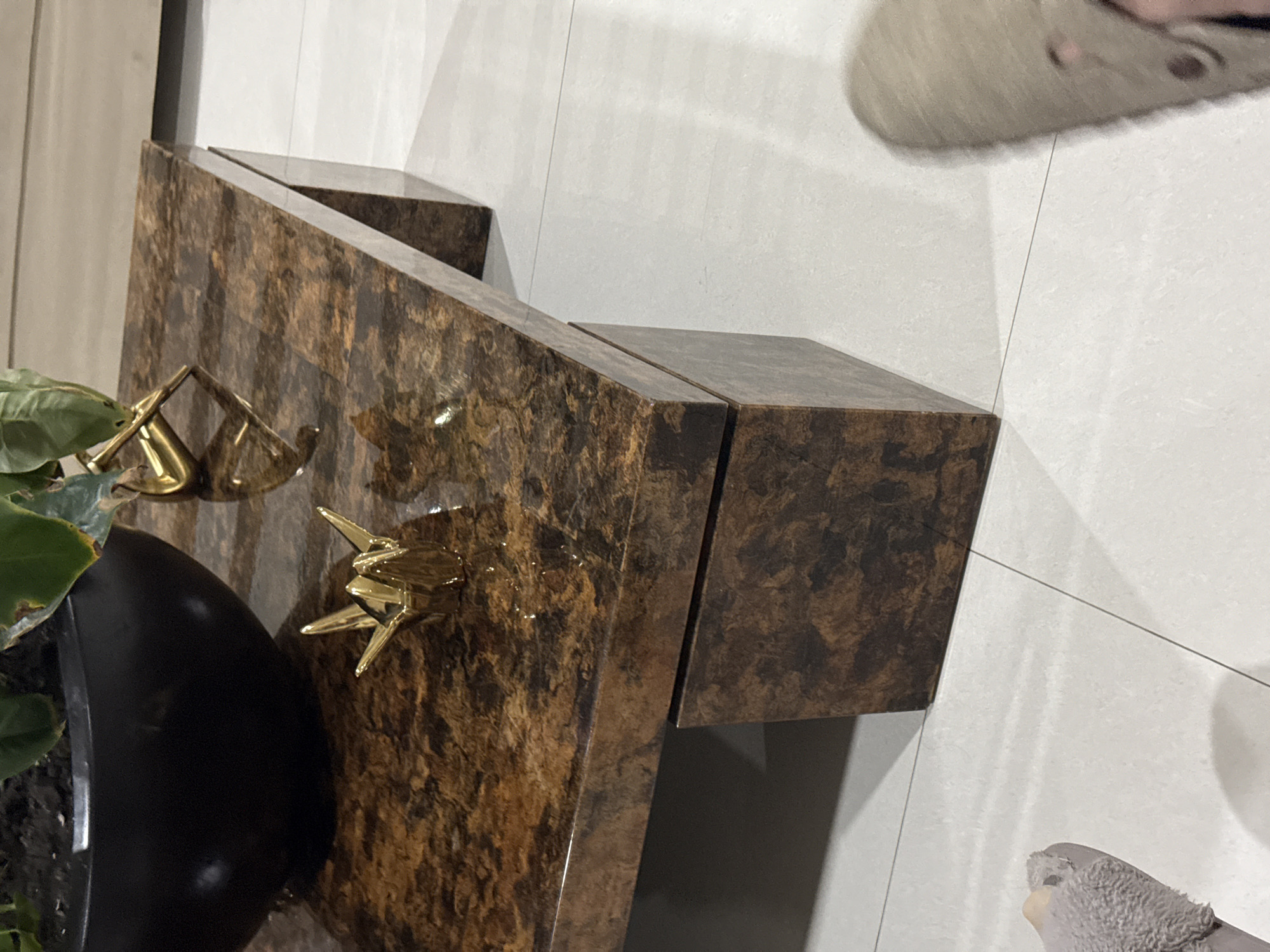 A high-angle, close-up shot shows a brown and black patterned countertop, with a black plant pot containing a green plant in the lower left. The countertop features two gold-colored decorative objects: one resembling a pair of scales and the other a multi-pointed star. The countertop appears to be installed against a white tiled wall. A beige slipper is visible in the upper right of the image and a grey slipper is visible in the bottom right. The location is not identifiable from the image alone.