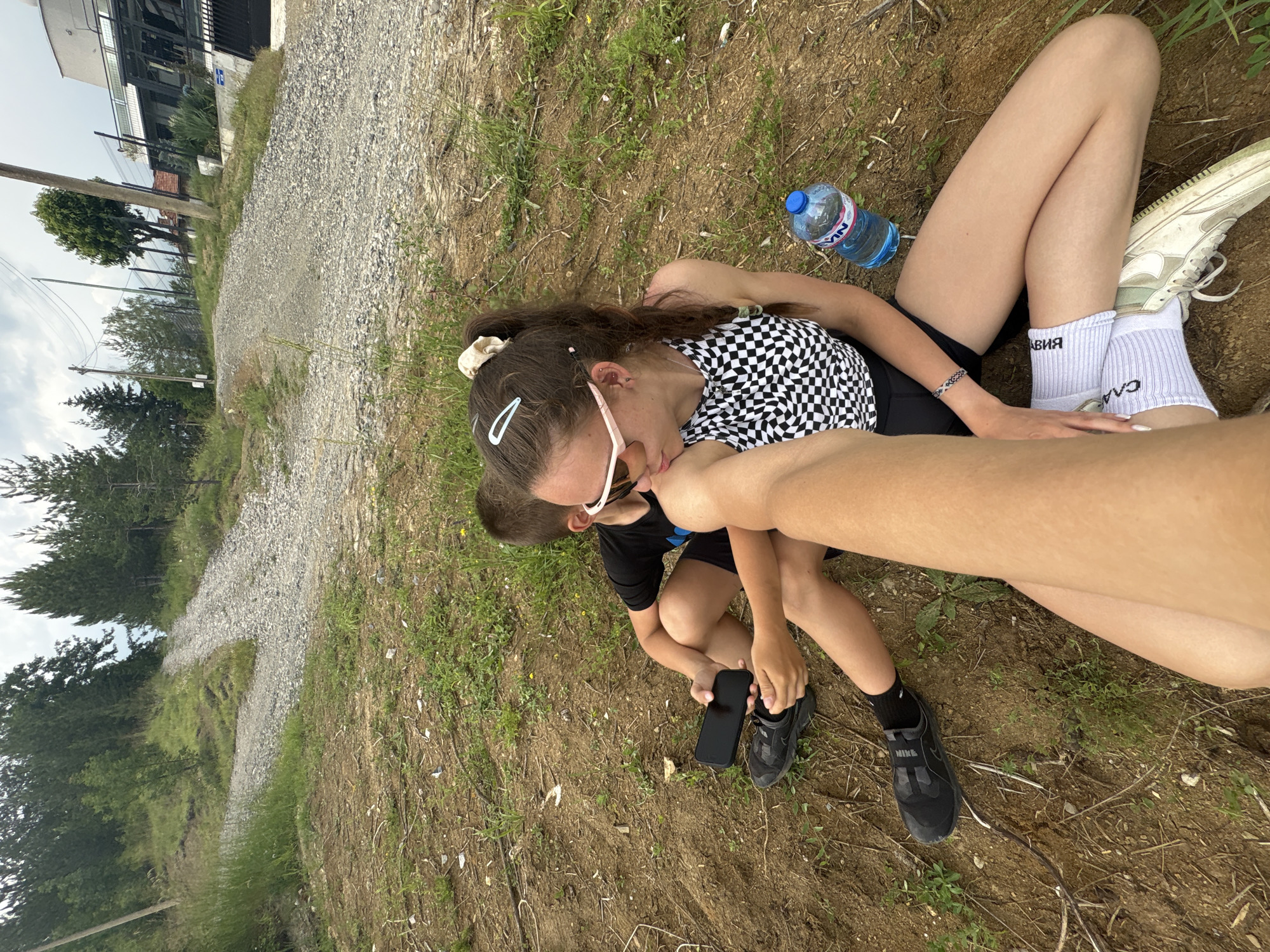 Two individuals are resting on the ground outdoors, next to a gravel path. The image is taken from a slightly elevated perspective, looking down on the subjects.
One person, positioned in the center, has long dark hair tied up with a light-colored scrunchie. They are wearing a black and white checkered top, black shorts, and white socks with black text that appears to say "АВИА" and "САА." They have light-colored sunglasses pushed up onto their head, light-colored hair clips, and earrings. Their left arm is visible, showing a braided bracelet. A bottle of water with a blue cap is resting on the ground near their head.
The other person, to the left and slightly below the first, is a younger individual with short brown hair. They are wearing a black t-shirt and black shorts, along with black sneakers and black socks. They are holding a black smartphone in their right hand.
The ground consists of dry dirt and sparse green grass. In the background, to the left, there's a gravel path winding through the scene. Beyond the path, there are several trees, including what looks like pine trees, and some utility poles. Further in the background, a building with a dark-colored fence or railing is visible, and the sky is overcast. The lighting suggests it is daytime. The overall mood appears relaxed, with the individuals resting.