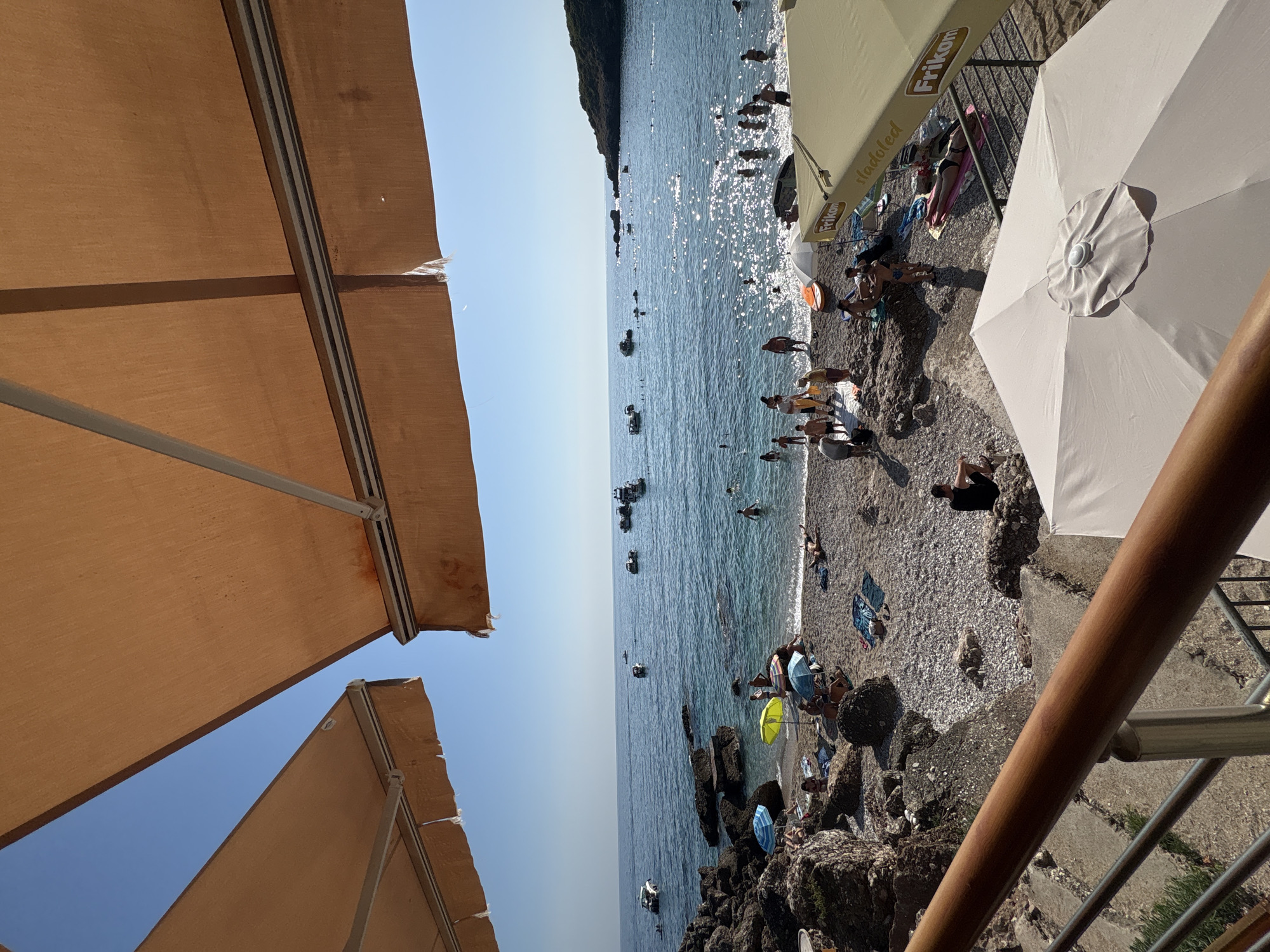 The image depicts a view of a crowded beach on a sunny day in Drobnići, Montenegro, as seen from beneath two canvas awnings. The awnings, one a light tan and the other a muted orange, partially obscure the clear blue sky above. The beach is composed of small grey and white pebbles, with large rocks scattered along the shoreline and in the water. Numerous people are present on the beach, some sunbathing on towels, others wading or swimming in the calm, clear blue water. Several colorful umbrellas, including a yellow and a blue one, provide shade. A few boats are anchored in the sea, with some individuals swimming around them. In the foreground, a wooden railing and metal balustrade are visible, suggesting a vantage point from a restaurant or cafe overlooking the beach. One light-colored umbrella is open in the foreground, and a cream-colored awning with the brand "Frikon" is partially visible to the right. The scene is vibrant with activity, indicating a popular tourist destination.