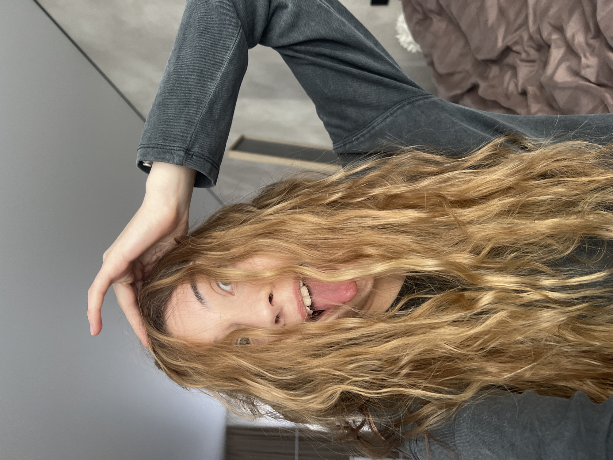 A young woman is upside down, sticking out her tongue. She has long, wavy, blonde hair. She is wearing a gray, long-sleeved shirt and has one hand on her head. The background shows a gray wall, a brown bedspread, and what appears to be flooring. Location: Unknown, Unknown.