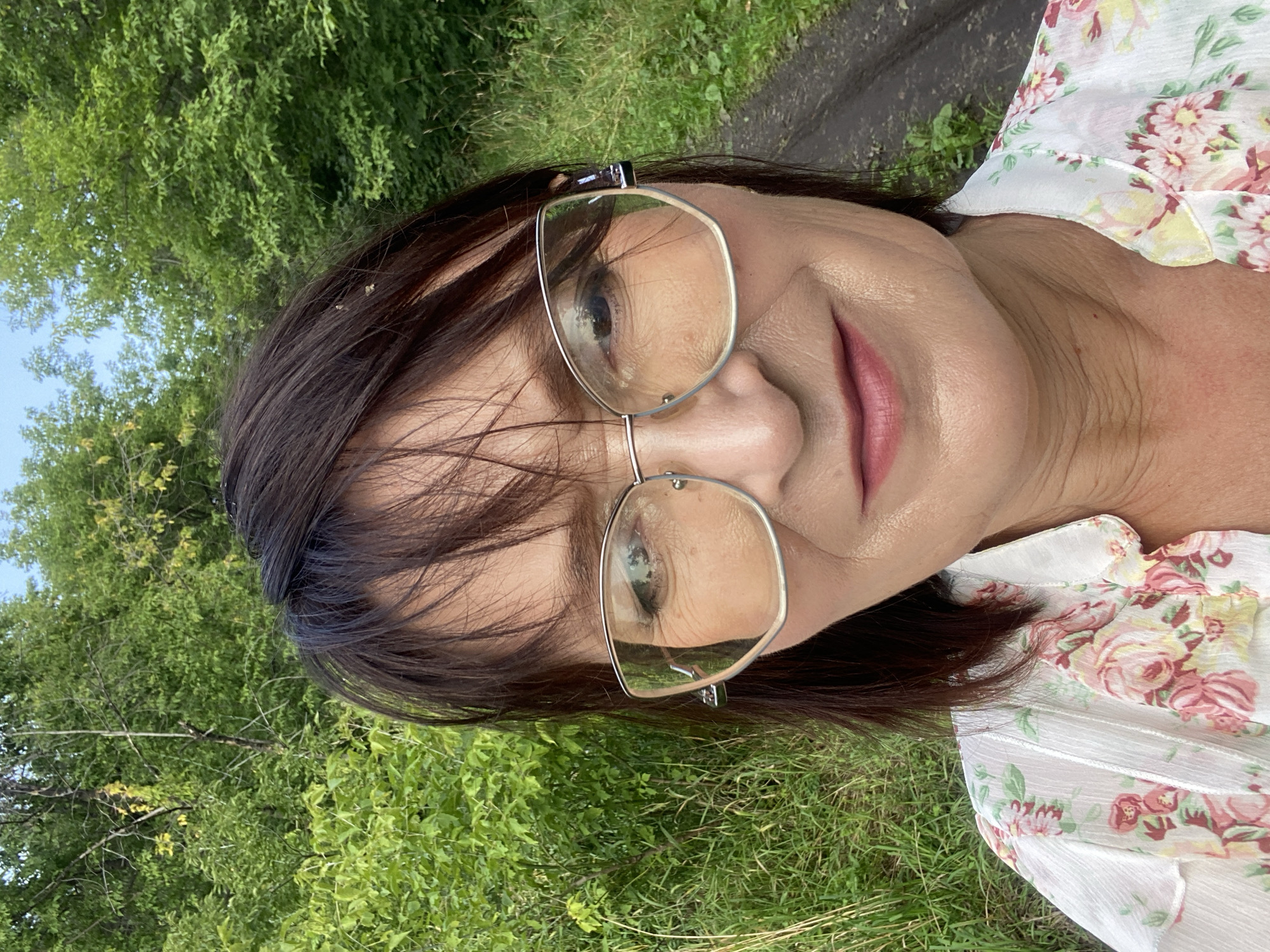 A medium shot selfie shows a woman with dark hair, wearing glasses and a floral blouse, with trees and greenery in the background. The scene appears to be a rural or park-like setting with a dirt path visible on the right. The woman is looking directly at the camera with a slight smile. The lighting suggests it is daytime. The location is specified as Novokuznetsk, Russia.