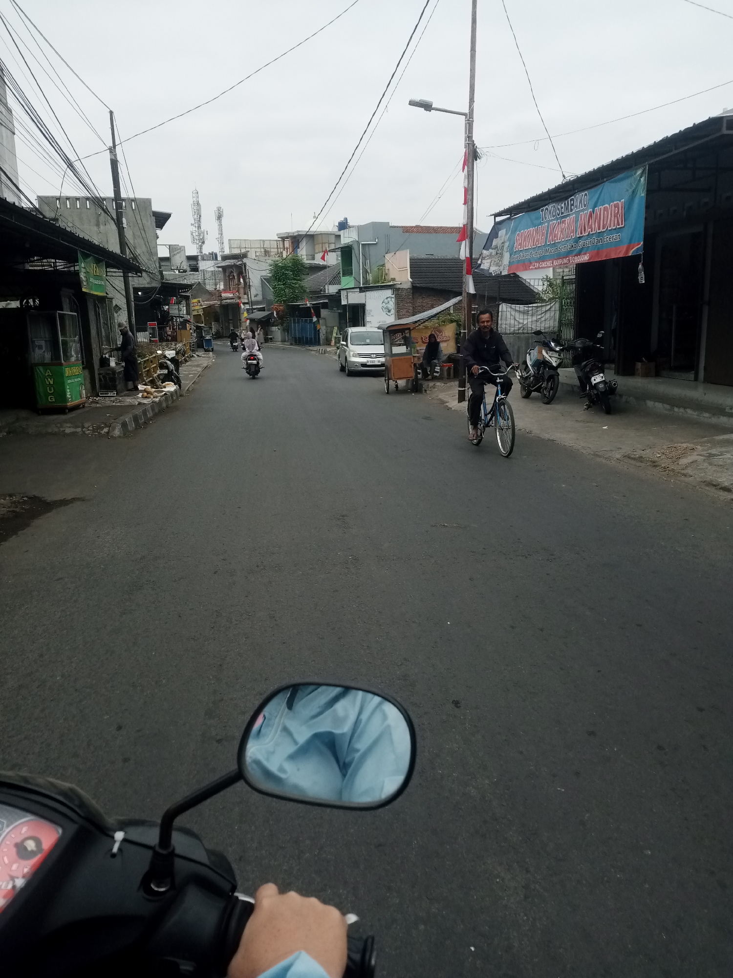 The image depicts a street scene in Tasikmalaya, Indonesia. In the foreground, a person's hand is visible on the handlebars of a motorcycle, with a mirror reflecting a light blue garment. The road is paved and appears to be a two-lane street.
Several motorcycles are parked along the side of the road, and two people are riding motorcycles further down the street. A man is riding a bicycle towards the camera, wearing a dark jacket and pants. On the right side of the street, there is a building with a blue banner that reads "TOKO SEMBAKO SAKINAH KARYA MANDIRI" and some Indonesian text below it. Various shops and stalls line both sides of the street, some with awnings.
In the background, buildings of various heights and construction styles are visible. Communication towers can be seen in the distance, indicating an urban or semi-urban environment. The sky is overcast. The overall impression is of a typical Indonesian street on a cloudy day, with ongoing daily activities like transportation and commerce.