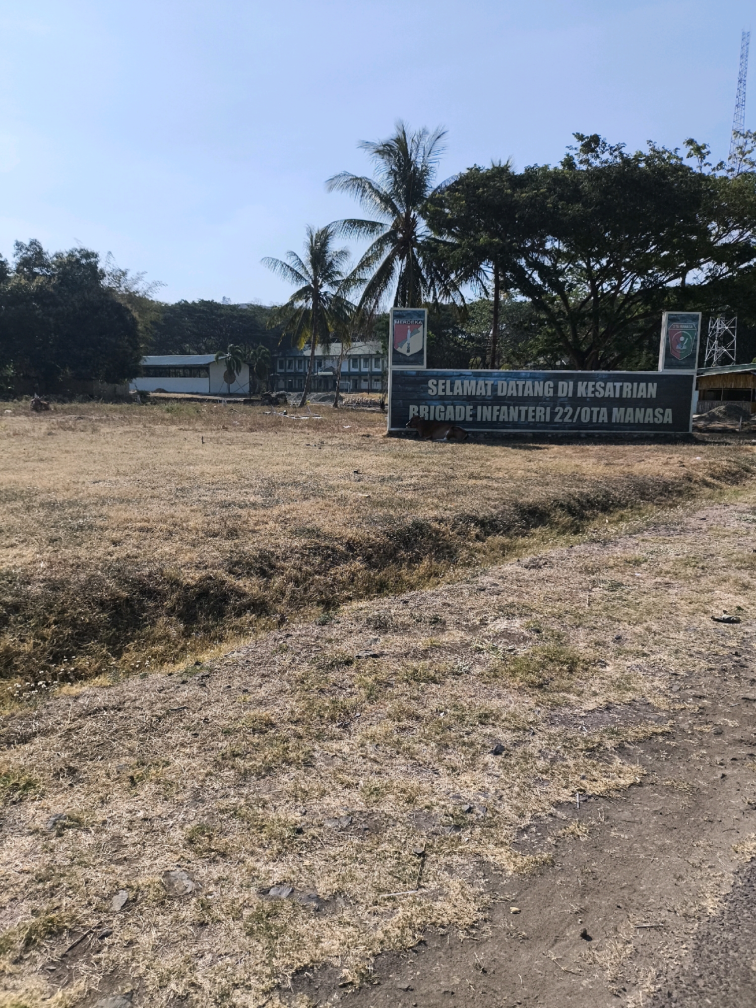 A wide shot captures a dry, grassy field in front of a building complex under a clear blue sky. In the foreground, the parched landscape is covered in light brown, dry grass and scattered stones, with a slight depression or ditch running diagonally across the middle. Behind the field, a long, dark sign with white text reads "SELAMAT DATANG DI KESATRIAN BRIGADE INFANTERI 22/OTA MANASA". Two crests are visible on the sign, one on the left with "MERDEKA" and another on the right with "OTA MANASA". To the left of the sign, a brown cow is lying down in the grass. Behind the sign, a multi-story building with white walls and green roofs is visible, surrounded by trees and vegetation. A cell tower is visible on the right side of the frame, peeking above the trees. The overall scene suggests a sunny, dry day in a military installation.