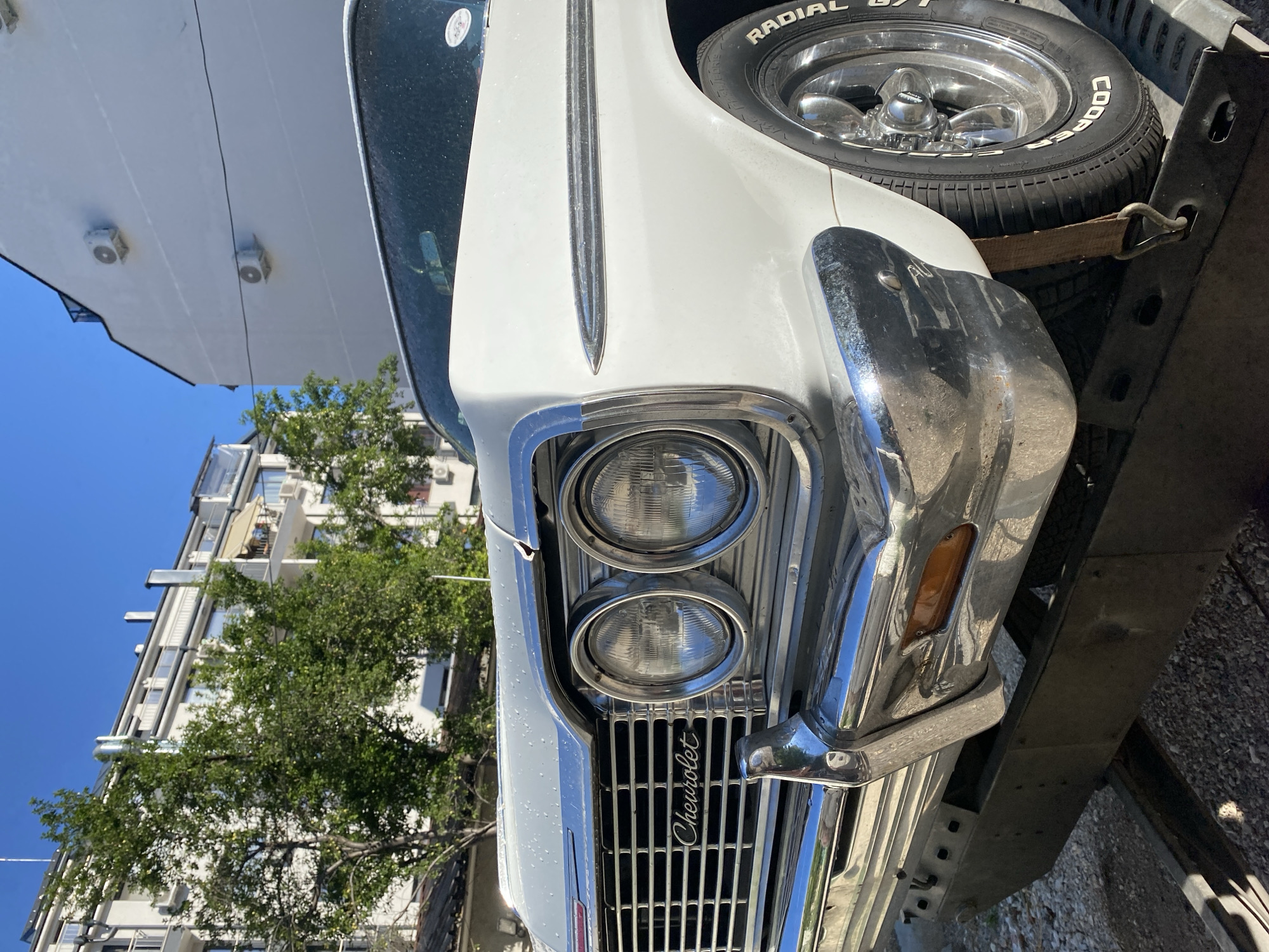 The image is a close-up, low-angle shot of the front of a white 1960s Chevrolet Impala parked on a trailer. The car's chrome grille, dual headlights with chrome bezels, and a portion of the chrome bumper are prominently displayed. The "Chevrolet" emblem is visible on the grille. A tire with "Cooper Radial" lettering is also in view. In the background, there is a white building with visible air conditioning units and trees with green foliage. The sky is clear and blue. The scene appears to be in an urban environment, possibly a street or parking area, with the car likely being transported. The specific location in the city of Pleven, Bulgaria, cannot be definitively determined from the visual information provided in the image. There are no people or discernible actions in the frame.