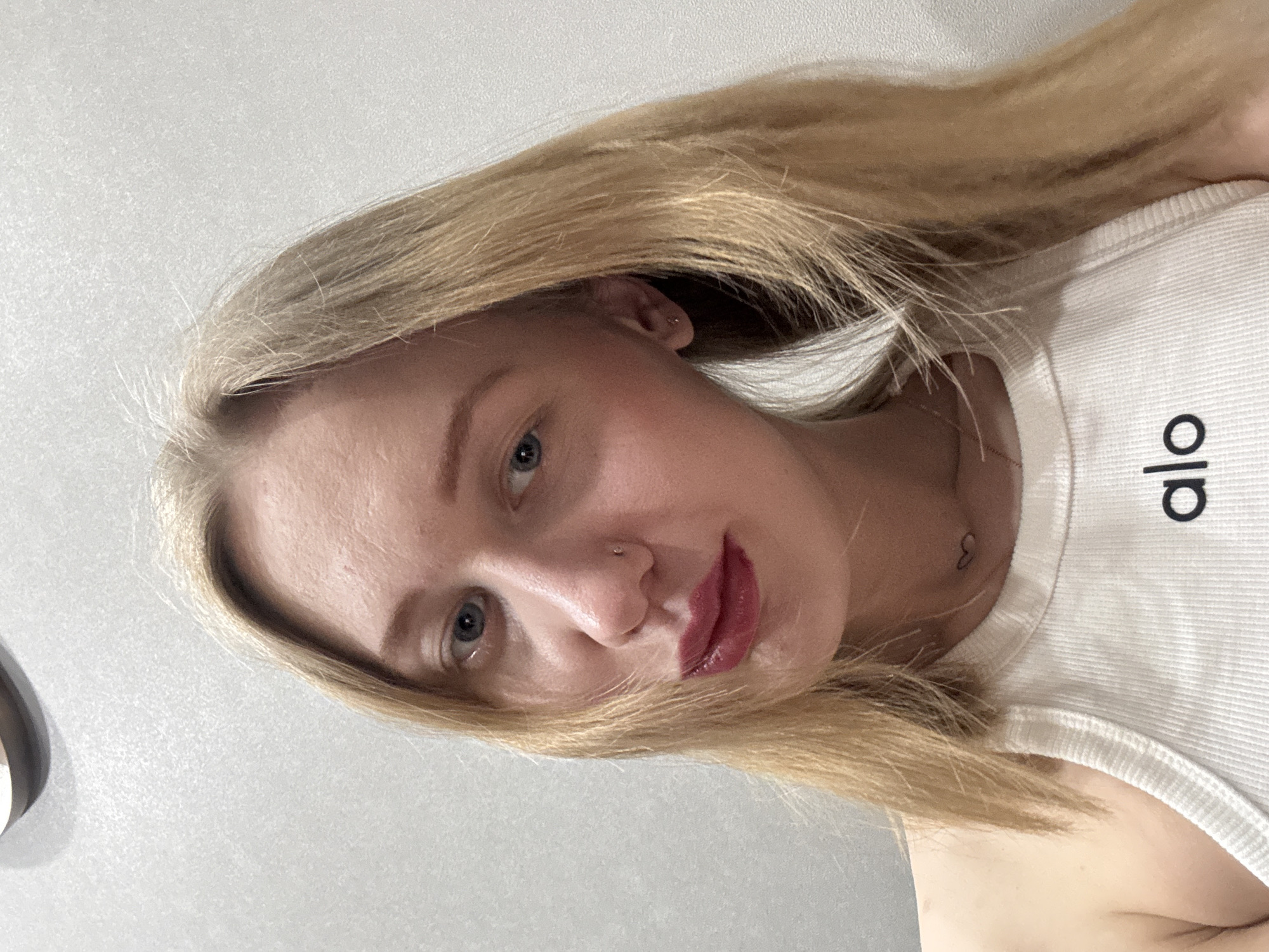 A blonde female is depicted in a close-up portrait, gazing directly at the viewer. She has blue eyes, dark red lipstick on her lips, and a small silver stud piercing in her left nostril. A similar stud is present in her left earlobe. A small, dark heart-shaped tattoo is visible on the left side of her neck, above a thin chain necklace. She is wearing a light-colored, ribbed tank top with the black letters "alo" printed across the chest. Her long, straight blonde hair frames her face and shoulders. The background consists of a plain, light-grey textured wall, with a segment of a light-colored, curved object visible in the upper left corner. The scene is located in the city of Novoaleksandrovka, Russia.