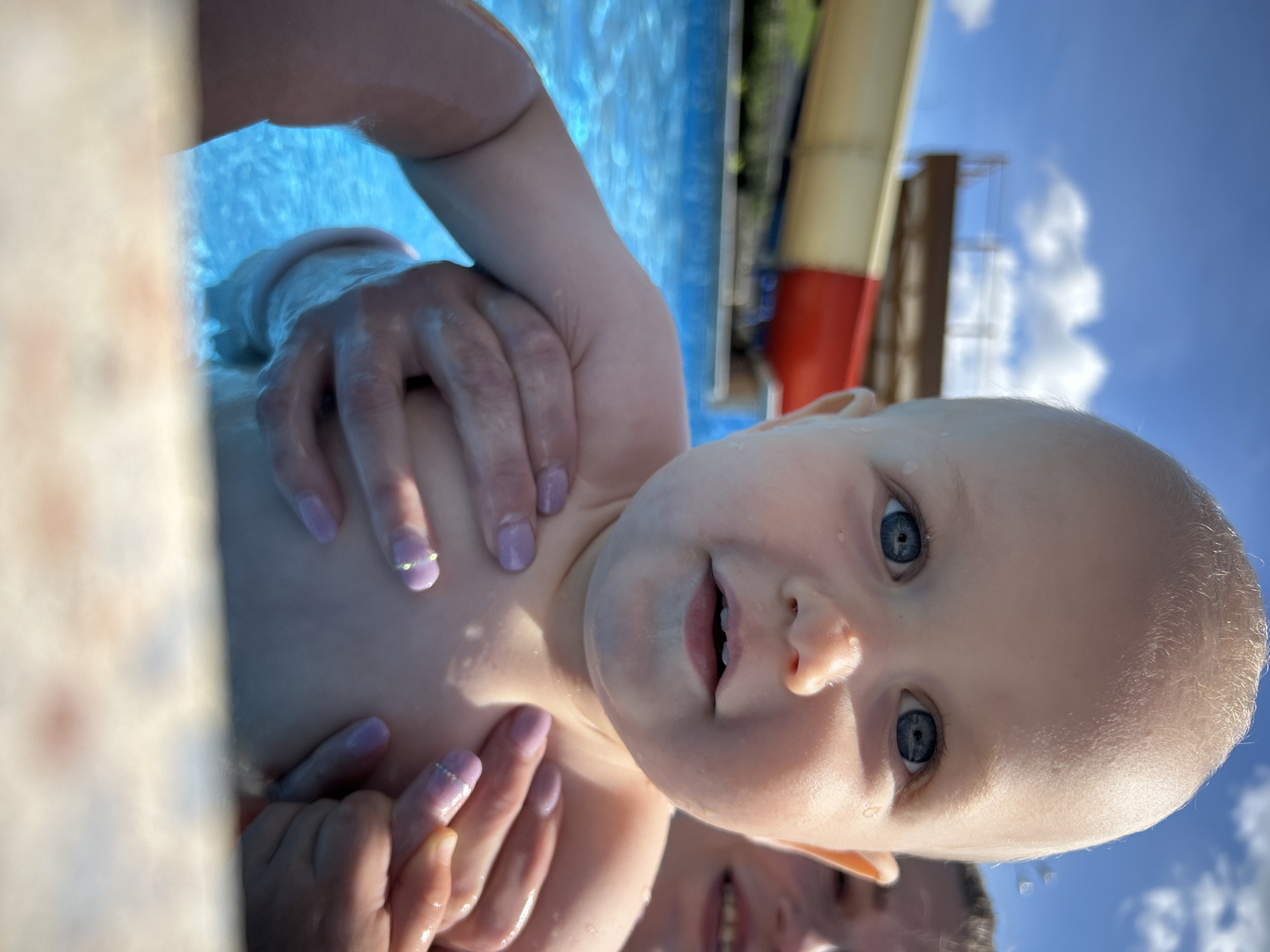 A baby with blue eyes is being held in a swimming pool by an adult whose hands are visible. The baby's head is close to the camera, showing a portion of their face. In the background, a water slide and blue sky with clouds are visible. The setting appears to be an outdoor swimming pool, likely in an area with recreational facilities. The city of Юбилейный, Russia is not explicitly depicted in the image, but the presence of a water slide suggests a public or private recreational area.