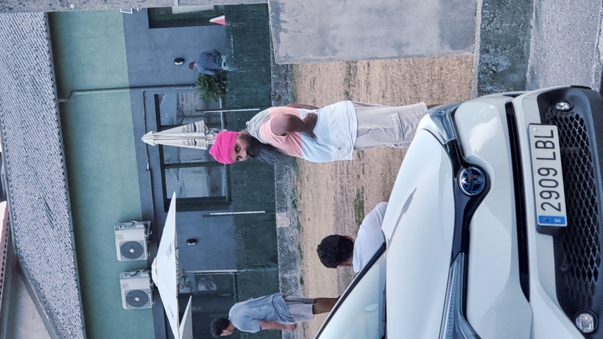 The image, taken in Sant Joan Les Fonts, Spain, depicts an outdoor scene with a focus on several individuals and a white car in the foreground. A man wearing a bright pink turban and a multi-colored t-shirt with light-colored cargo shorts stands with his back partially to the camera, looking towards the viewer. He has a beard and is leaning on his hip. To the left, another man in a light blue t-shirt and light-colored shorts is seen from the back, also looking away. In the middle ground, a third person is seated, facing away, wearing a light-colored shirt. Behind them, a building with a grey tiled roof and a greenish-grey facade is visible. Two air conditioning units are mounted on the upper part of the building. Several umbrellas, one white and one folded and beige, are situated near the building. A portion of a white car with a license plate reading "2909 LBL" is prominent in the lower part of the frame. The ground is covered in dirt and gravel. A concrete wall runs along the right side of the image.