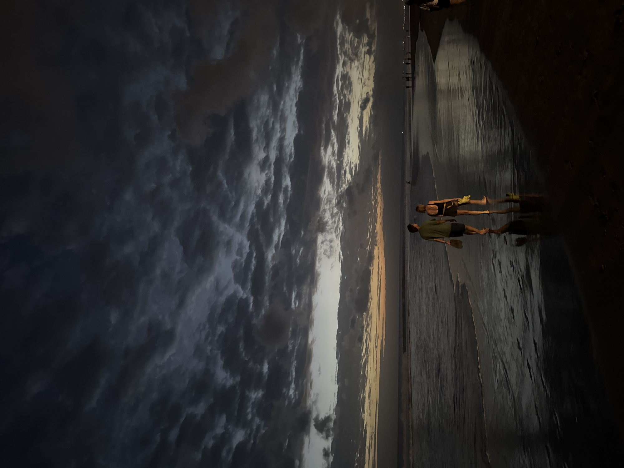 The image shows a beach scene at dusk, likely in Kuta, Indonesia, given the characteristic dark clouds and the appearance of the ocean and sand. Two people, a man and a woman, are walking away from the camera on the wet sand near the water's edge. The man is wearing a green t-shirt and dark shorts, and the woman is wearing a black tank top and shorts, carrying a yellow object. They are silhouetted against the darkening sky, which is filled with dramatic, textured clouds that obscure the setting sun, although a faint orange glow is visible on the horizon. The ocean is calm with gentle waves lapping at the shore, reflecting the dim light. Further down the beach, other individuals and groups are visible as small, distant figures. The overall atmosphere is serene and somewhat moody, characteristic of a tropical beach at twilight.