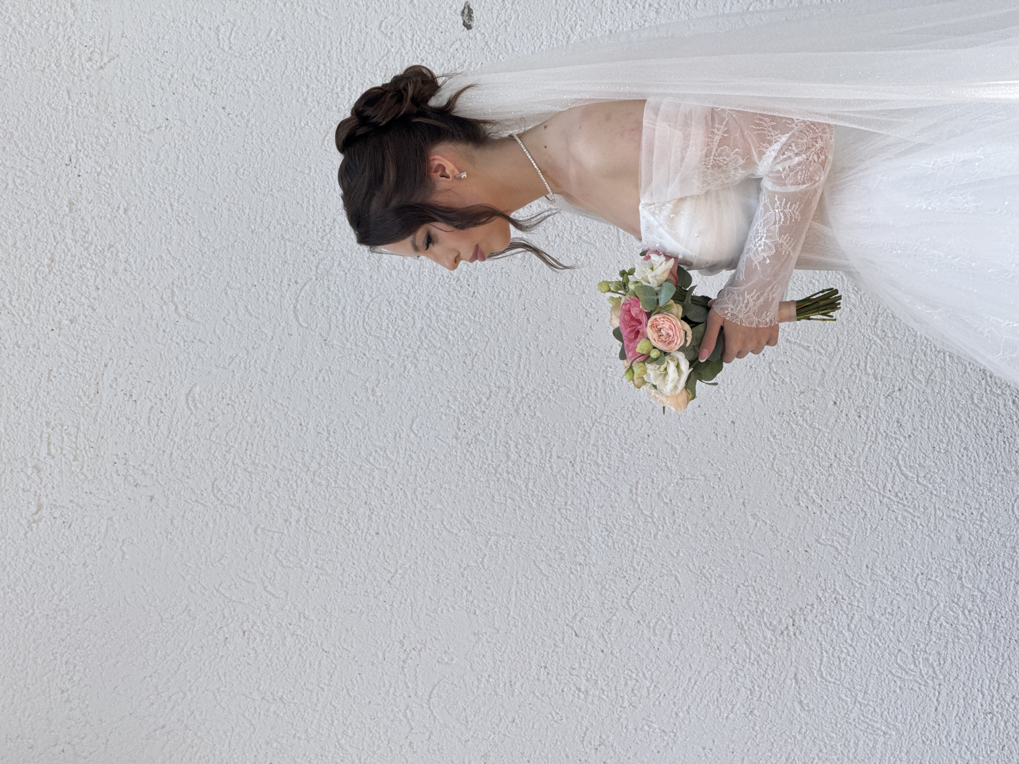 A bride in a white wedding dress with an off-the-shoulder neckline and long lace sleeves stands in profile against a textured white wall. She holds a bouquet of pink and white flowers. A sheer veil with glitter detail cascades down her back. Her hair is styled in an updo, and she wears a delicate pearl necklace and stud earrings. The scene appears to be outdoors, possibly in the city of Votkinsk, Russia, given the architectural style of the wall. The lighting suggests daytime.