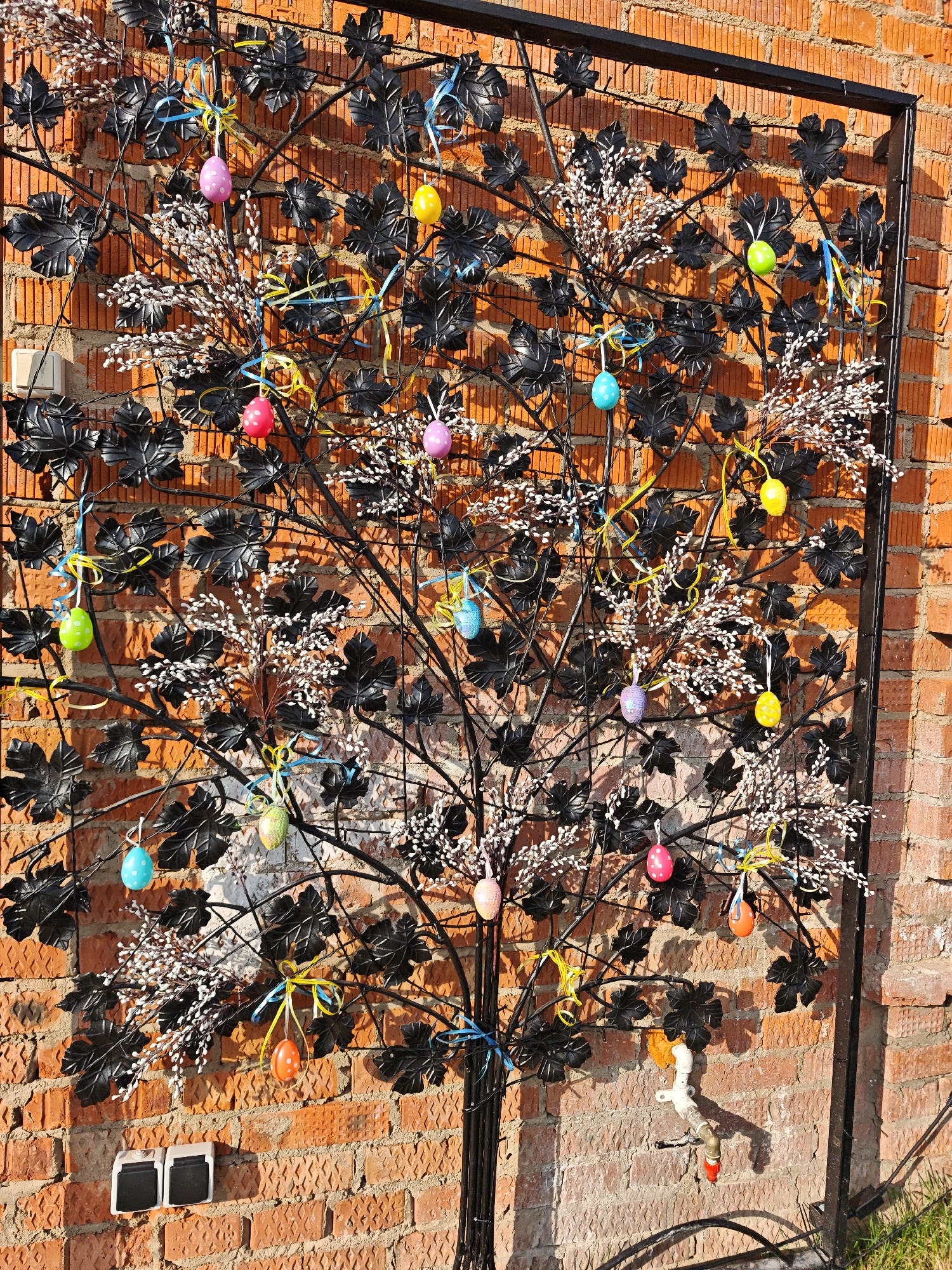 A metal art piece resembling a tree is mounted against a red brick wall. The "tree" has black metal leaves and white bud-like decorations. Several colorful Easter eggs hang from the branches with ribbons. A water faucet and two electrical boxes are also visible on the brick wall beneath the tree. The photo was taken in Жестылево, Russia.