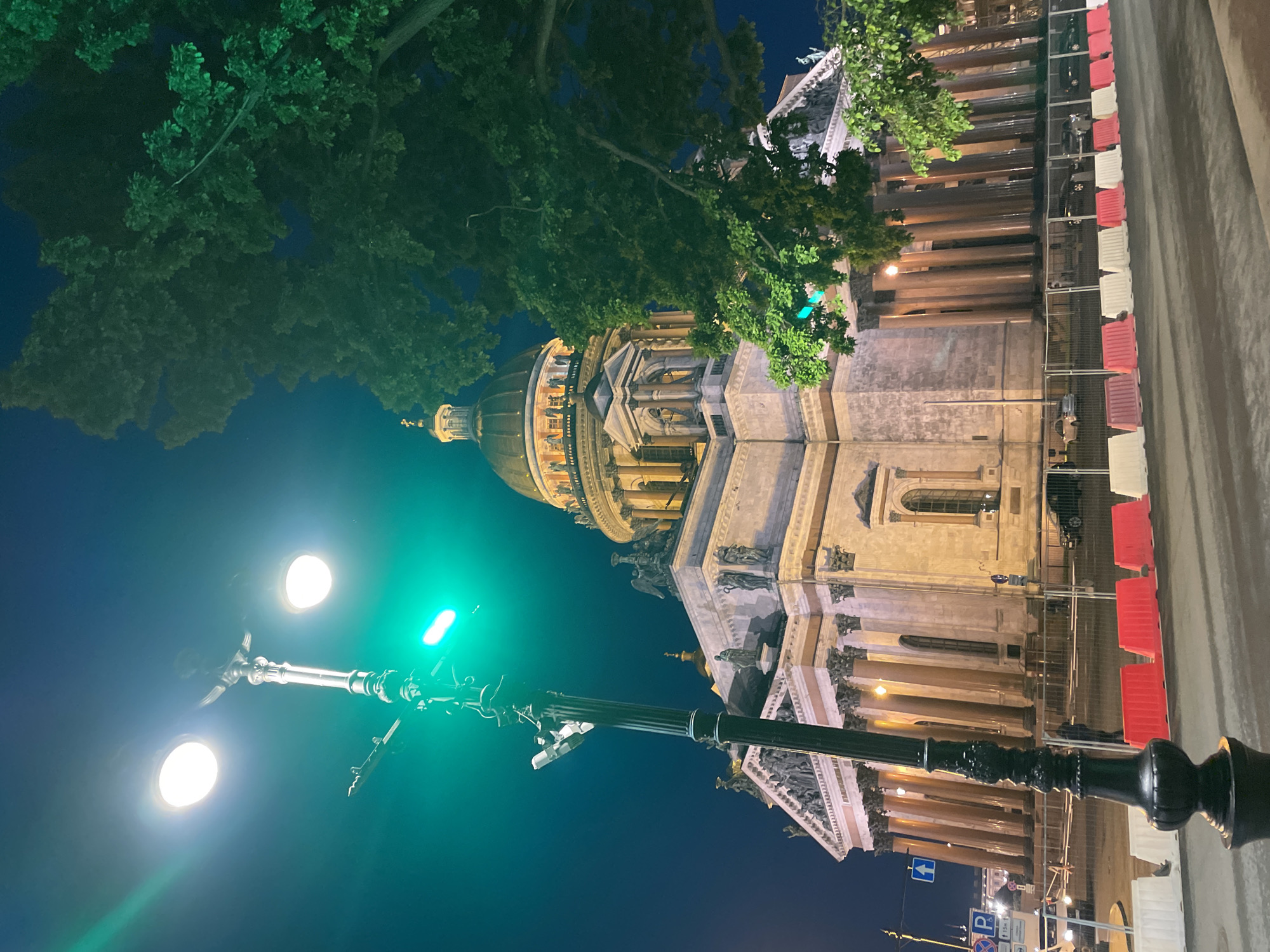 This night-time image captures a grand, classical-style building, which is Saint Isaac's Cathedral in Saint Petersburg, Russia, viewed from a low angle looking upwards. The massive structure features a prominent golden dome, illuminated by artificial lights, and is supported by numerous large columns. Ornate friezes and sculptures are visible along the upper parts of the building, showcasing intricate architectural details.
In the foreground, a decorative black streetlight pole stands tall, equipped with two bright white lights and a striking green light that casts a distinct glow onto the scene. Lush green tree foliage partially frames the building and the dark night sky, adding a natural element to the urban setting.
Along what appears to be a street or pathway to the right of the building, red and white plastic road barriers are lined up. Behind these barriers, some dark-colored vehicles are faintly visible. The scene is illuminated by artificial light sources, indicating it is nighttime. The sky is dark and clear. Visible road signs include a blue sign with a white upward-pointing arrow and another blue sign with a white 'P' symbol, indicating parking. No people or specific events are depicted, conveying a serene and majestic atmosphere.