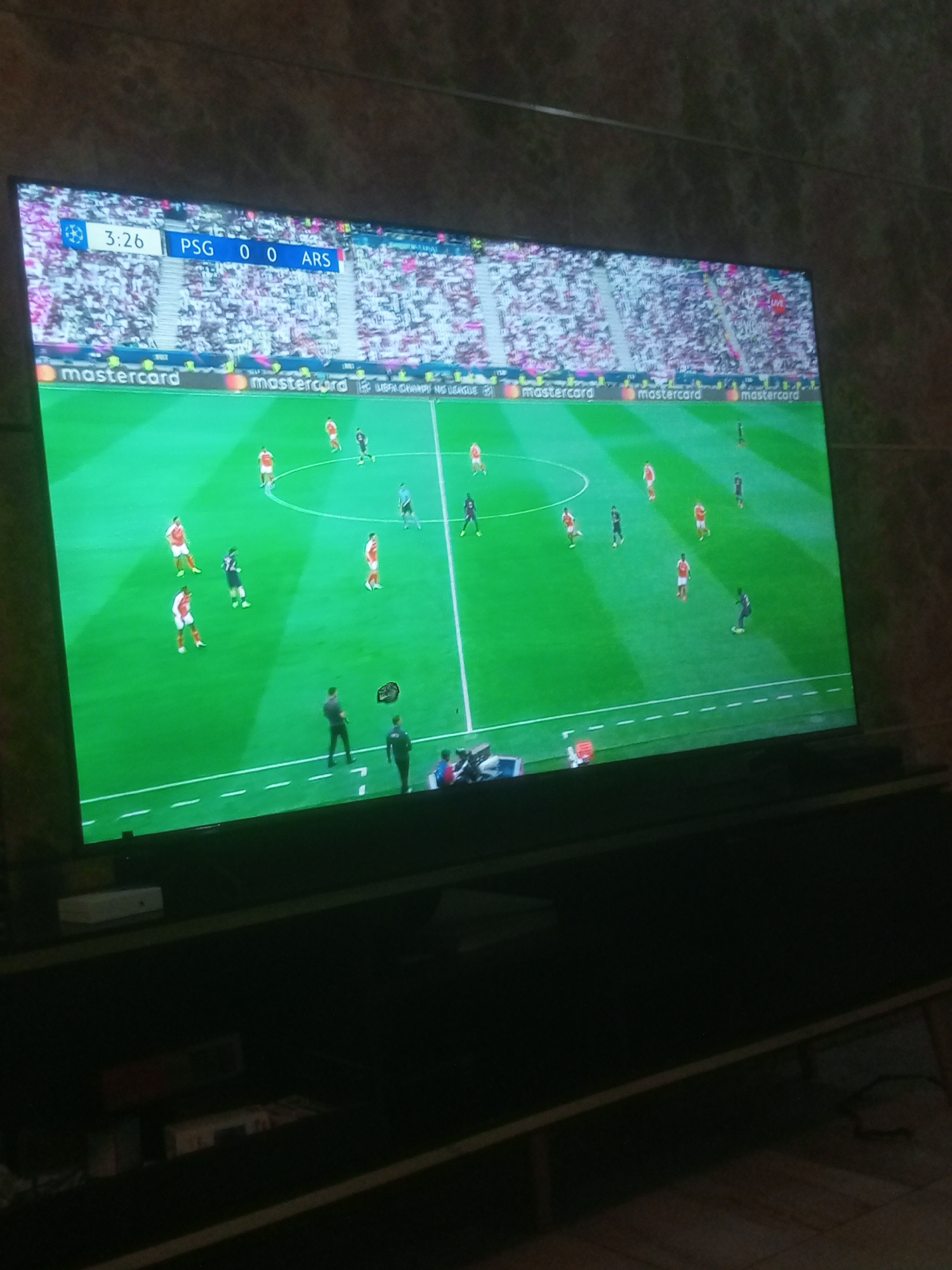 The image shows a television displaying a soccer game between Paris Saint-Germain (PSG) and Arsenal (ARS). The score is 0-0, and the game is 3 minutes and 26 seconds in. The game appears to be a UEFA Champions League match, judging by the branding displayed along the top of the screen. A partial view of the stadium, filled with spectators, can be seen in the background of the broadcast. The television is placed on a dark-colored stand. The location is Unknown, Unknown.
