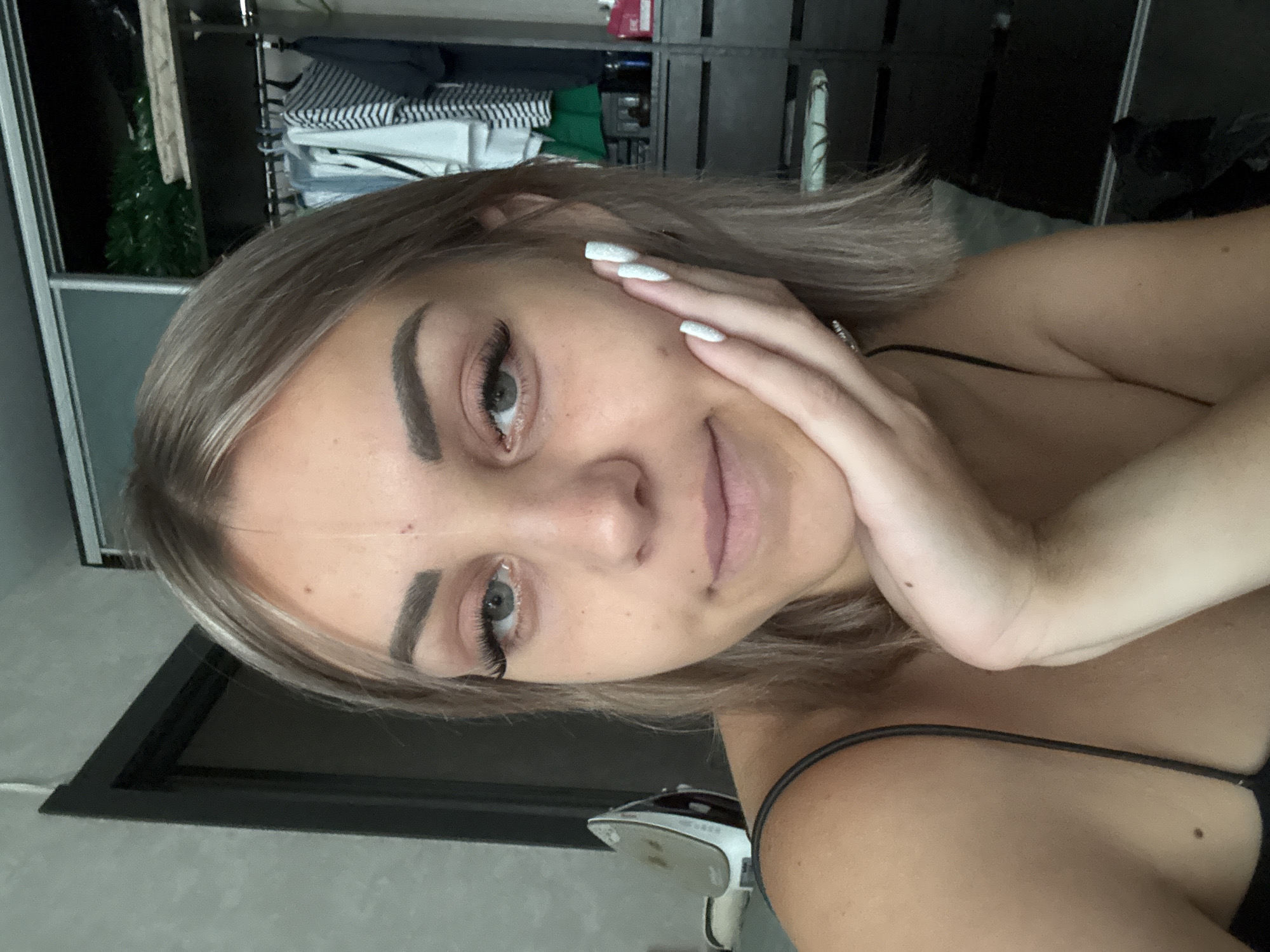 A young woman with blonde hair and blue eyes is pictured in a medium close-up, her right hand resting on her cheek. She is wearing a black spaghetti strap top. Her nails are painted white and appear to have glitter. In the background, to the left, a grey wall and a dark door frame are visible, with a silver and white steam iron on a surface. To the right, a closet with sliding doors is partially open, revealing shelves with clothing, including striped shirts and green items. The image appears to be taken indoors, possibly in a residential setting in Yuzhno-Sakhalinsk, Russia.