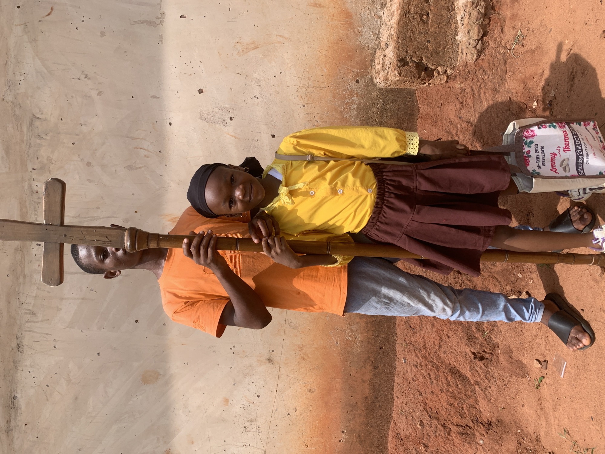 A young man and a girl, presumably siblings, are standing shoulder-to-shoulder, holding a wooden cross/staff. The man is wearing an orange T-shirt and light blue jeans, while the girl is wearing a yellow top, a brown skirt, and a black head covering. The girl is holding a tote bag with the text "DE-IVKE 2023 (OBEREMPA) Ammy weds Ikenna Saturday 21st OCT 2023 CELEBRATION OF LOVE". They are positioned against a wall and a dirt ground. The context suggests a location in Ukwu-Abwa, Nigeria.