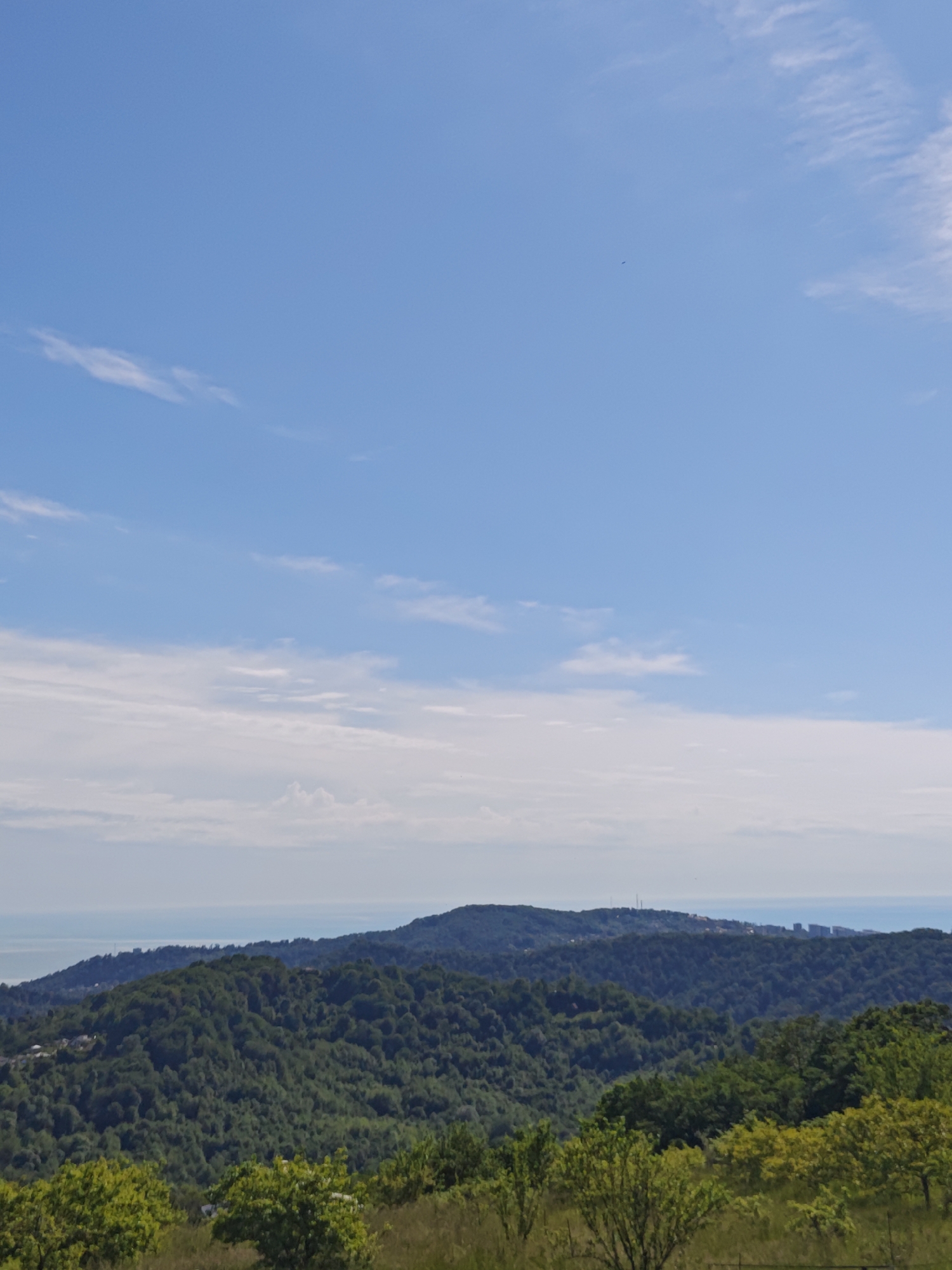 The image depicts a landscape in Прогресс, Russia. In the foreground, there are trees and grassy terrain. Moving towards the midground, a series of lush, green forested hills rise, with one taller hill in the center. In the background, the sea is visible, with a horizon line indicating a body of water. Above the sea, the sky transitions from a light blue near the horizon to a deeper blue overhead, with scattered white clouds.