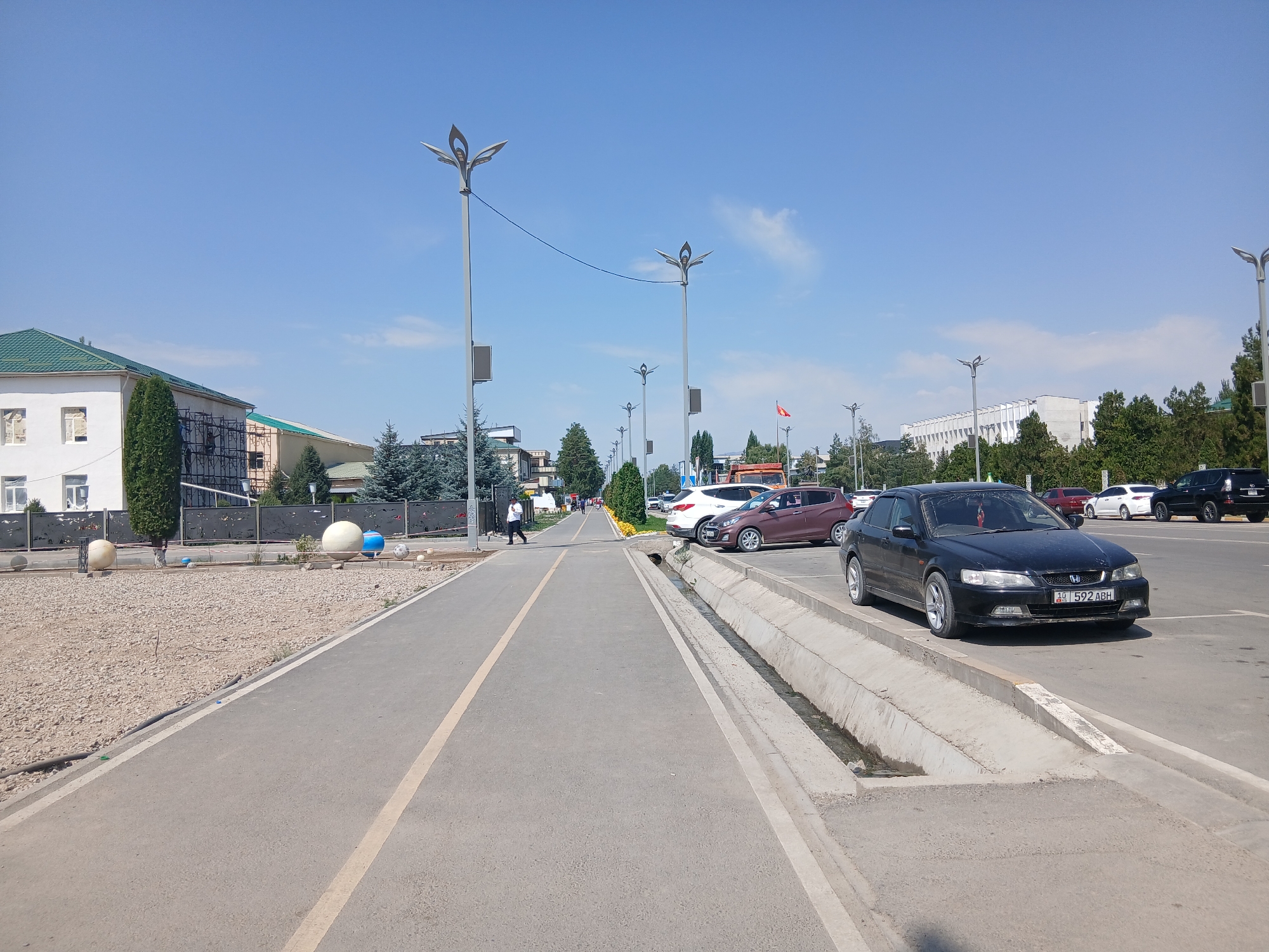 The image depicts a street scene in Talas, Kyrgyzstan, on a clear, sunny day. A paved pedestrian walkway runs along the left side of the frame, bordered by a gravel area with large spherical decorative elements. A dark sedan is parked on the right side of the street, adjacent to a concrete drainage ditch. Several other vehicles are visible parked further down the street, along with streetlights and trees. A lone pedestrian is walking on the walkway in the mid-ground. In the distance, buildings and more trees line the street. A flag, possibly the flag of Kyrgyzstan, is visible on a flagpole to the right. The overall environment appears to be an urban or semi-urban setting, with a focus on public infrastructure like walkways and roads.