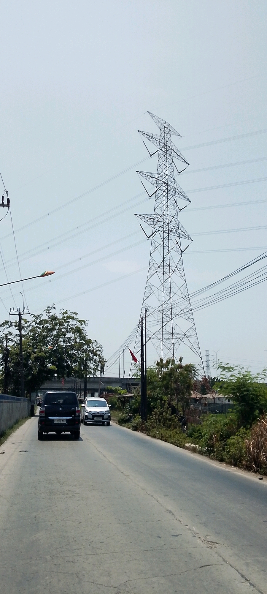 The image depicts a road scene in Srimahi, Indonesia, characterized by a bright, overcast sky. Dominating the right side of the frame is a tall, lattice-style electricity pylon, a prominent feature of the industrial or infrastructure landscape. Multiple power lines stretch across the sky, originating from the pylon and extending towards the left. A lamppost with a colored head is visible on the left side, situated near utility poles and more power lines.
The road itself is paved and appears somewhat worn, with visible cracks and uneven patches. A black SUV is in the foreground, driving away from the viewer in the left lane. To its right and slightly ahead, a white car is also traveling in the same direction. The road is flanked by vegetation, with a cluster of trees on the left and denser foliage, including tall grasses and shrubs, on the right. In the background, there is evidence of structures and possibly agricultural land, hinting at the semi-urban or semi-rural nature of the location. A small red flag is attached to a pole near the electricity pylon. The overall impression is one of a typical day in a developing area with significant electrical infrastructure.