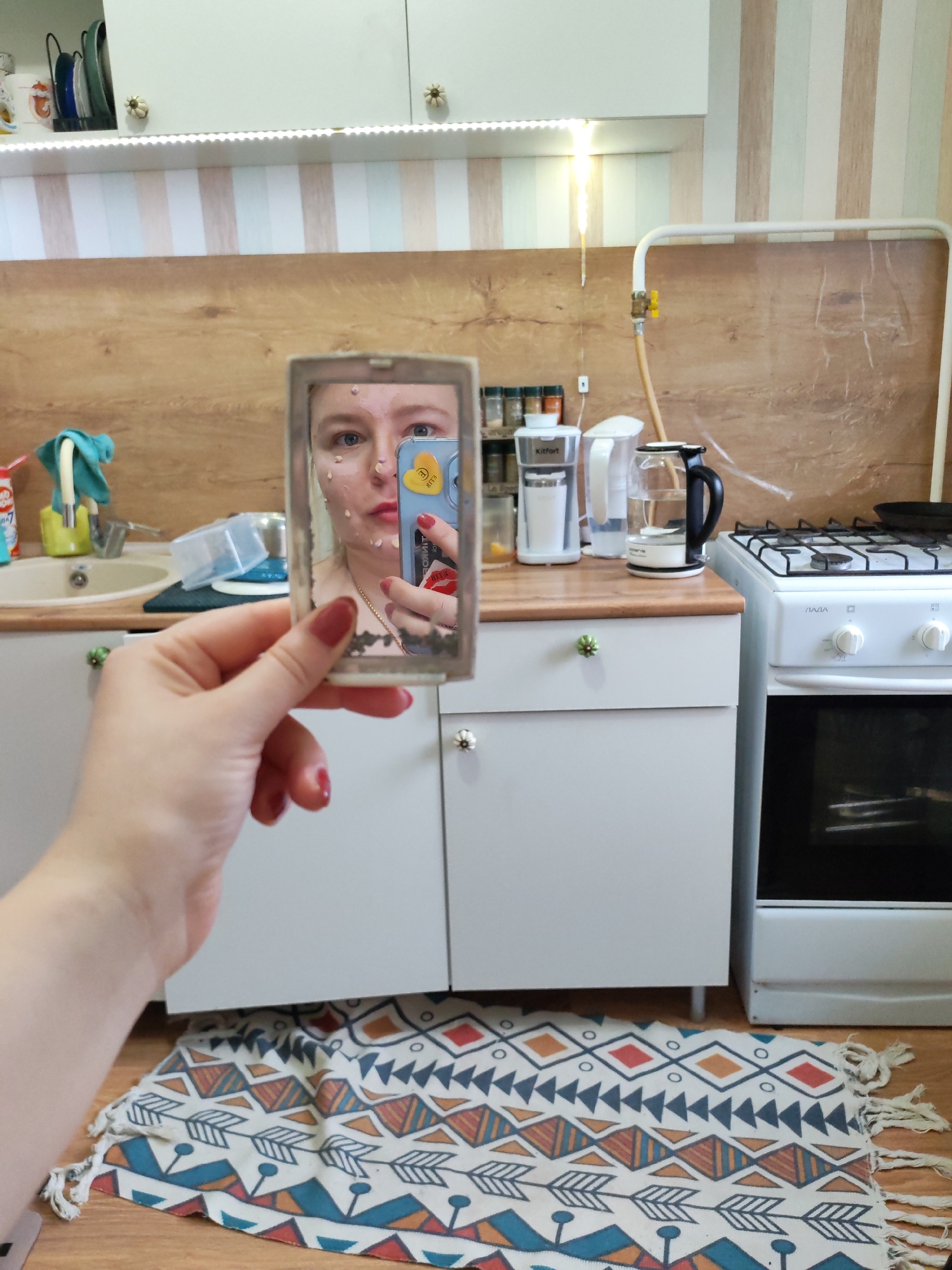 A person is holding a small mirror in their left hand, reflecting their face with what appears to be cosmetic dots on the skin. The person is also holding a blue smartphone with a yellow heart-shaped sticker on the back. The background shows a kitchen setting, including kitchen cabinets with flower-shaped knobs, a sink, a stove, a kettle, and a coffee maker. There is a patterned rug on the floor. Based on the Cyrillic text "ЛАДА" visible on the stove, and the style of the kitchen, the location is Izhevsk, Russia.
