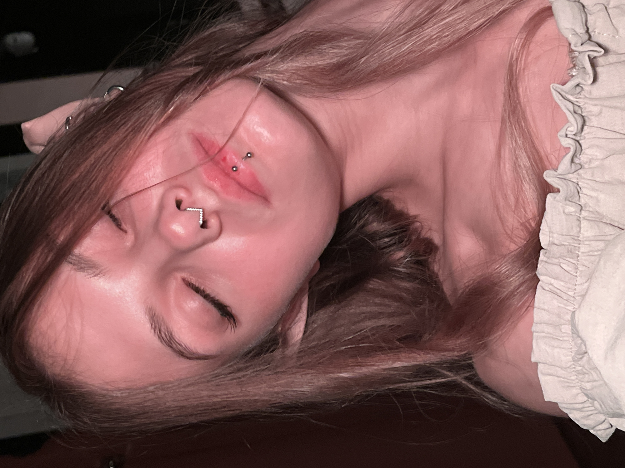 A close-up, tilted image of a young woman lying down with her eyes closed. She has light-colored, medium-length hair and fair skin. She has a septum piercing and piercings on her lip. She wears a light-colored garment with a ruffled edge. The background is dark and out of focus. There is no indication of the location being in Фоки, Russia.