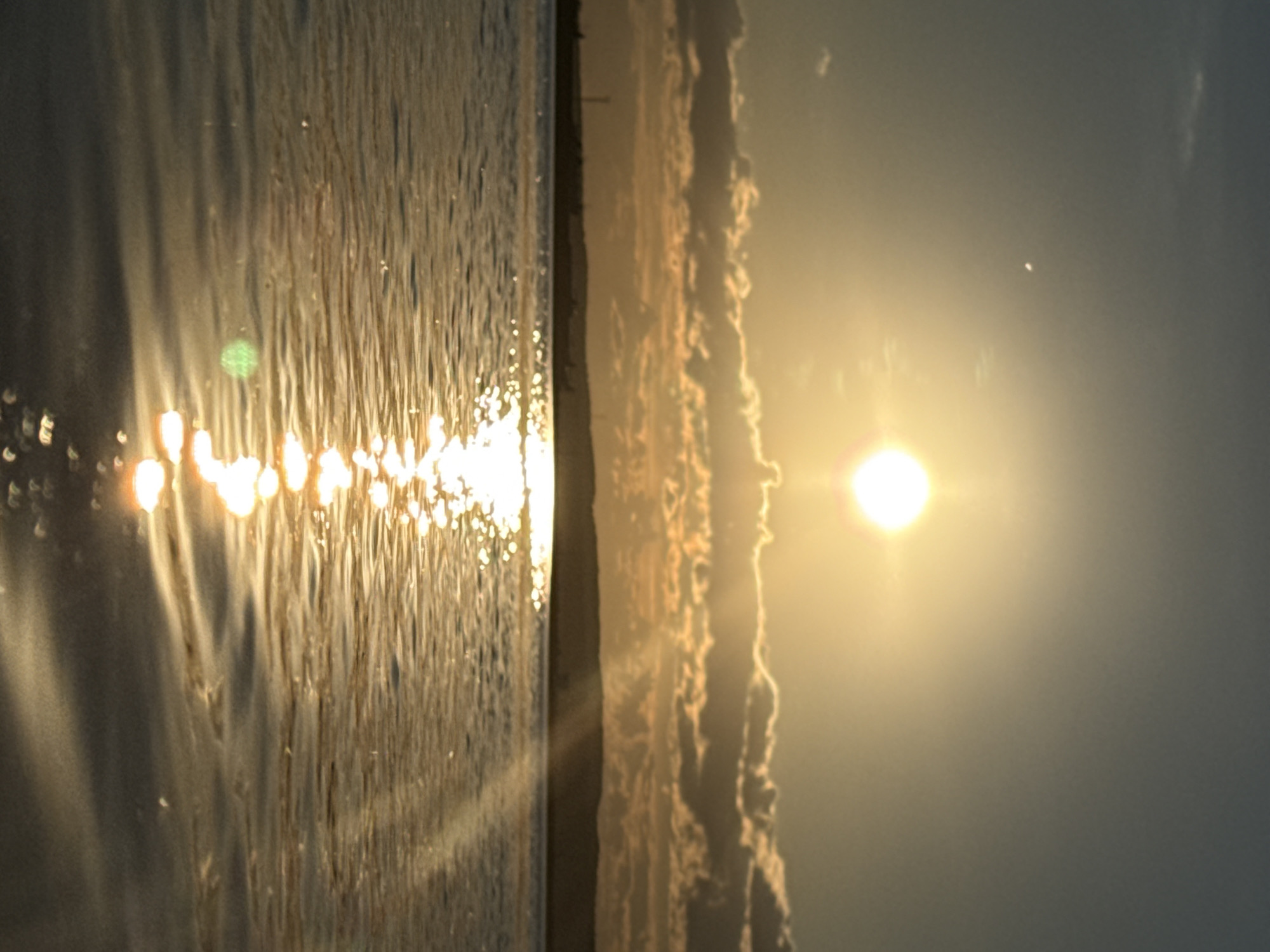 The image captures a vibrant sunrise or sunset over a body of water, likely the Kama River or Votkinsk Reservoir in Chaikovsky, Russia. The scene is bathed in a warm, golden light.
On the right side of the frame, a bright, circular sun glows intensely, low above the horizon. Its powerful light casts a brilliant golden hue across the entire sky and reflects dramatically on the water's surface.
The left side of the image is dominated by the body of water, which appears to have gentle ripples or small waves. The direct sunlight creates numerous shimmering reflections and sparkling highlights on the water, appearing as bright, elongated streaks and dots of light. Several lens flares are visible within this area, including a distinct green circular flare.
A distinct horizontal line separates the water from the distant horizon. This horizon features an irregular, textured band, which could be a distant shoreline, landmass, or a formation of low-lying clouds, all partially silhouetted and backlit by the intense golden light.
The sky above the horizon transitions from a bright golden around the sun to a softer, muted blue-gray further away. A small, bright white dot is visible high in the sky, positioned above and slightly to the left of the sun. The overall atmosphere conveyed is serene and peaceful. There is no visible text in the image.