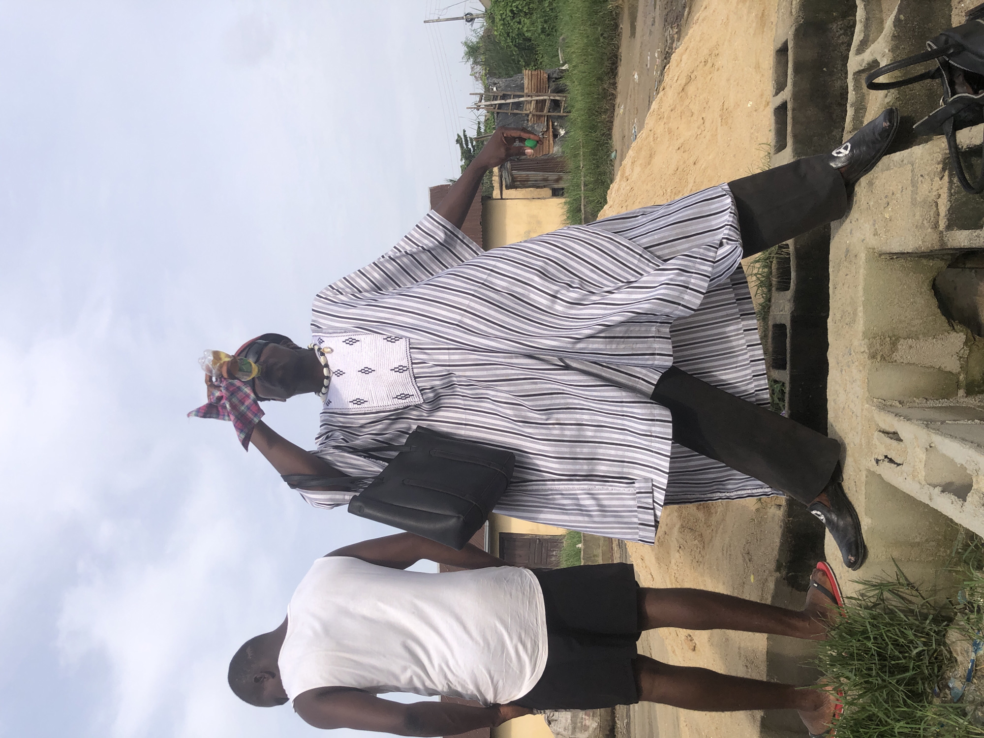 In Ado, Nigeria, an adult stands beside a person in a white tank top and black shorts. The adult is wearing a striped robe, dark pants, and dress shoes, with a black bag on his shoulder. He holds a small green object in one hand and a cloth in the other. They are near concrete structures and greenery. The background includes buildings and utility poles under a cloudy sky.