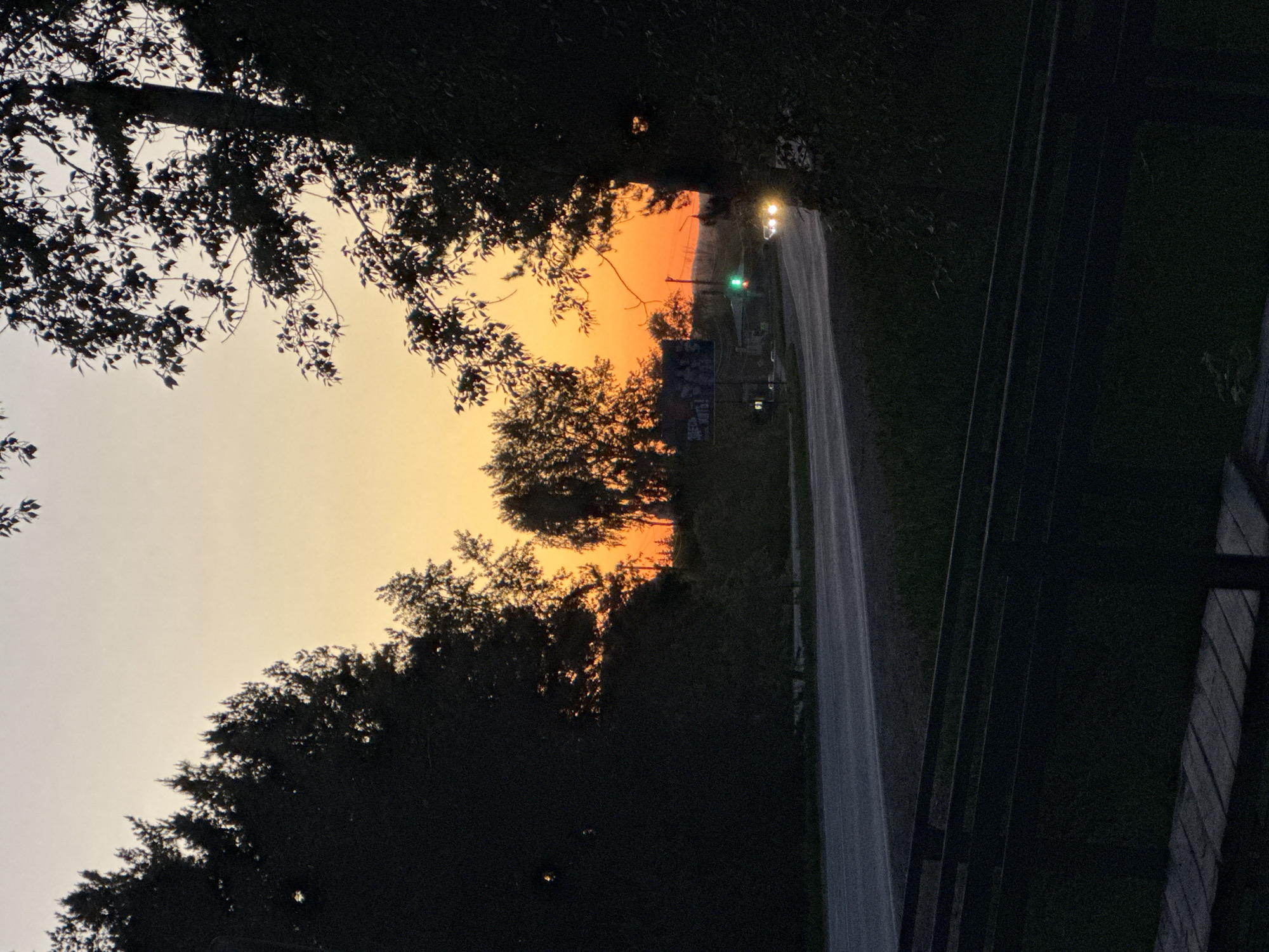 The image captures a sunset scene on a road. A two-lane asphalt road stretches into the distance, illuminated by the headlights of a vehicle and a green traffic light. The sunset paints the sky with orange and yellow hues, partially obscured by silhouettes of trees lining the roadside. A building with a visible sign is situated along the road. Fencing is also visible on the side of the road. The location is not specified, and no information regarding Berezovka, Russia, is contained in the image.