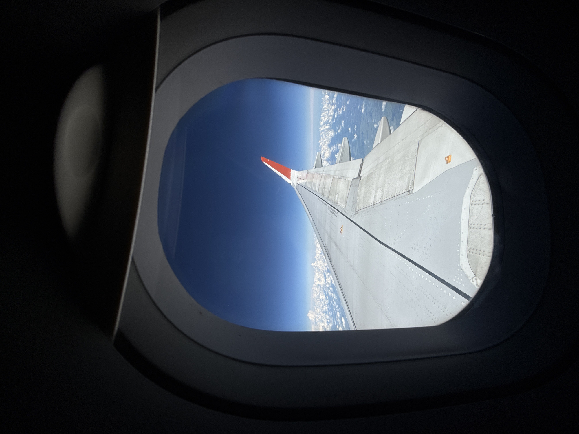 This image is a view from an airplane window, looking out at the wing and clouds. The sky is a deep blue, and the clouds are white and fluffy. The wing is white with a red tip. There are no people or any indication of the city of Усть-Ламенка, Russia. The scene is serene and vast, suggesting a high altitude flight.